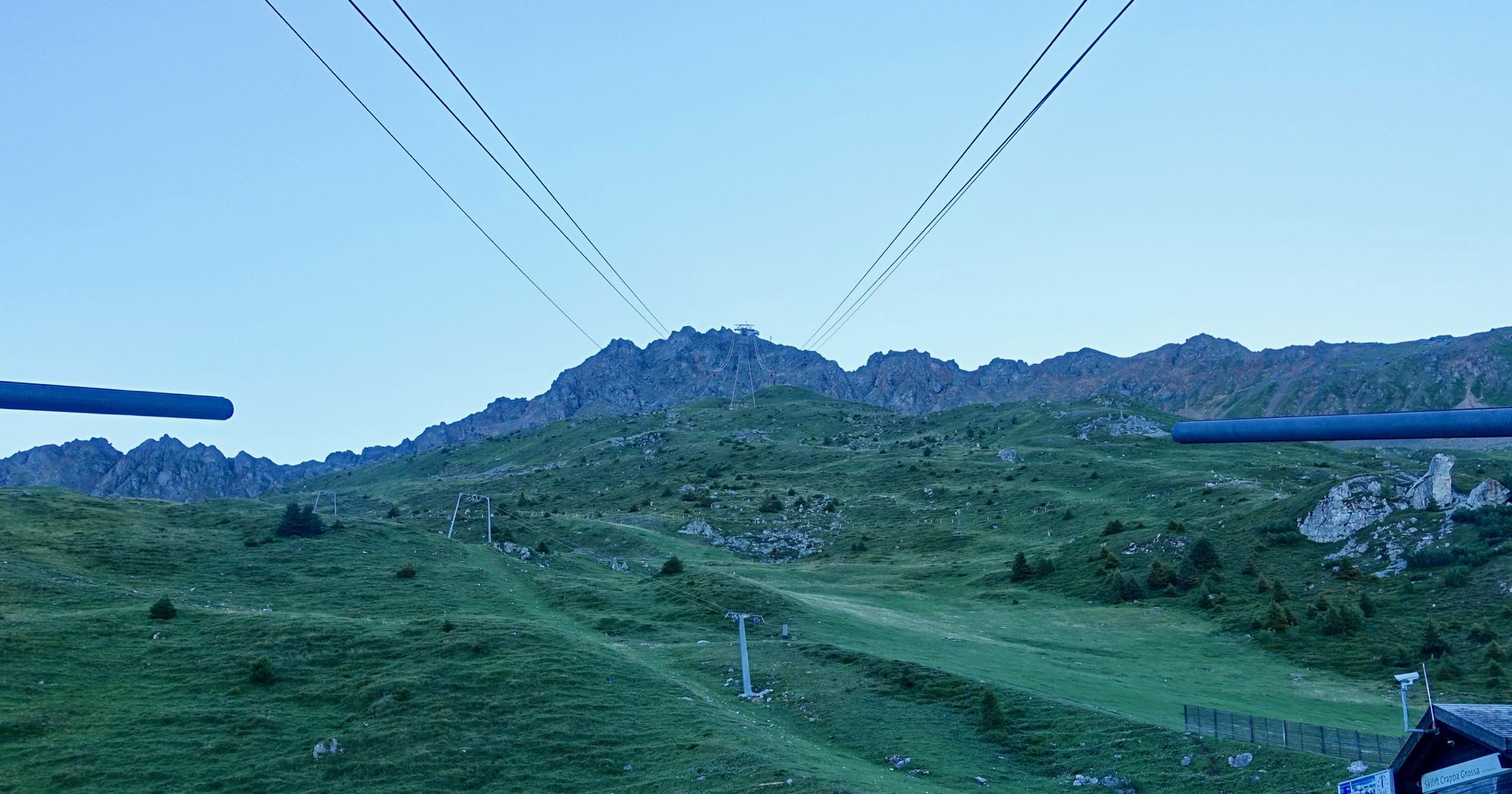 View from a ski lift looking towards a mountainous landscape, featuring grassy slopes and rocky peaks under a clear blue sky. The image includes cable lines leading toward the distant mountain summit. Rothorn Zone mountain bike trail.