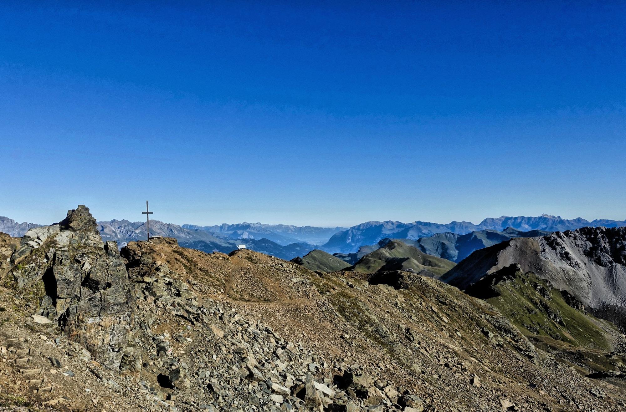 A rocky mountain landscape under a clear blue sky, featuring a cross on a stone outcrop with distant mountain ranges in the background. Rothorn Zone mountain bike trail.