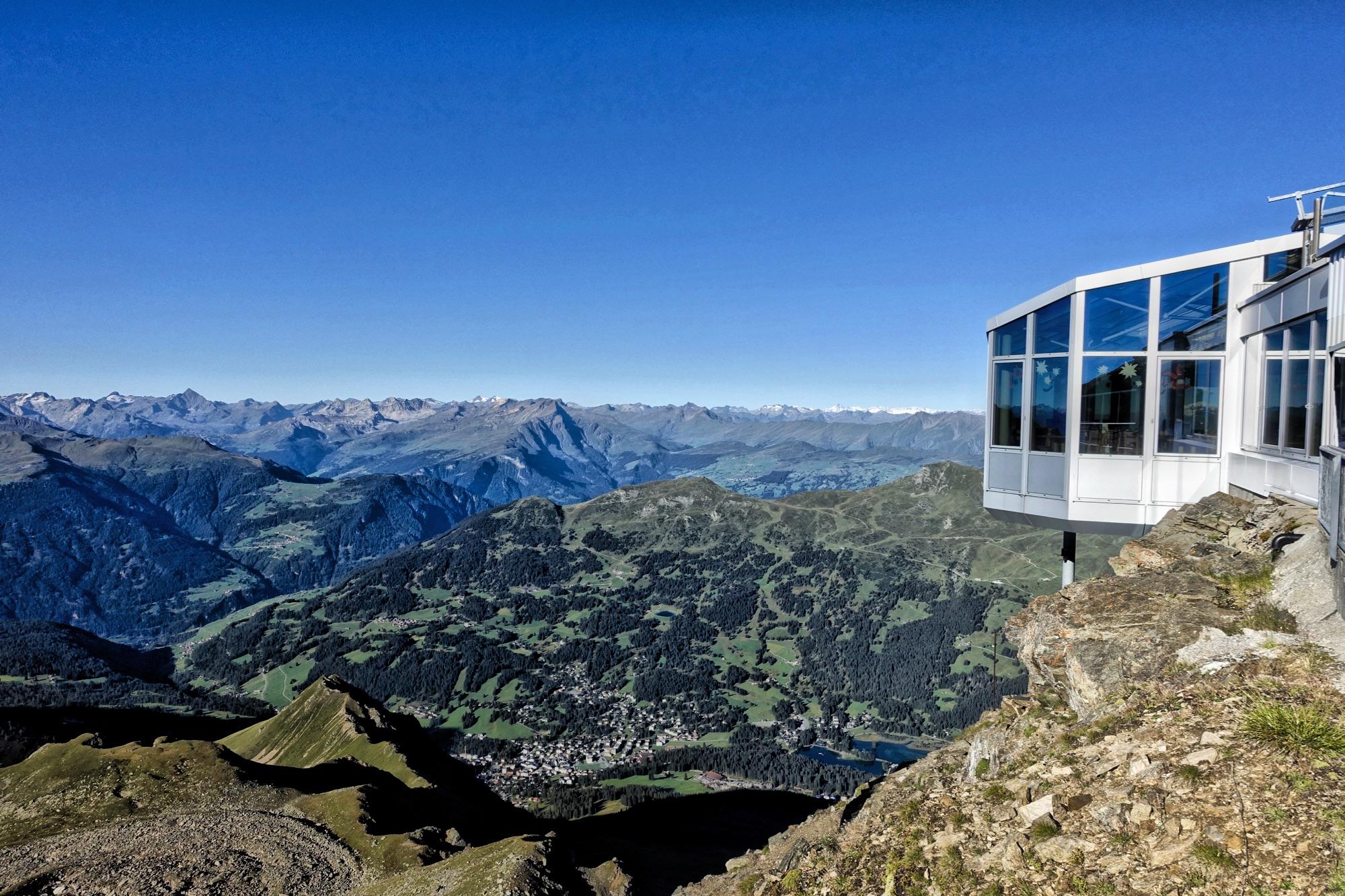 A panoramic view of a mountainous landscape under a clear blue sky, featuring lush green hills and distant snow-capped peaks, with a modern glass structure perched on a rocky ledge in the foreground. Rothorn Zone mountain bike trail.