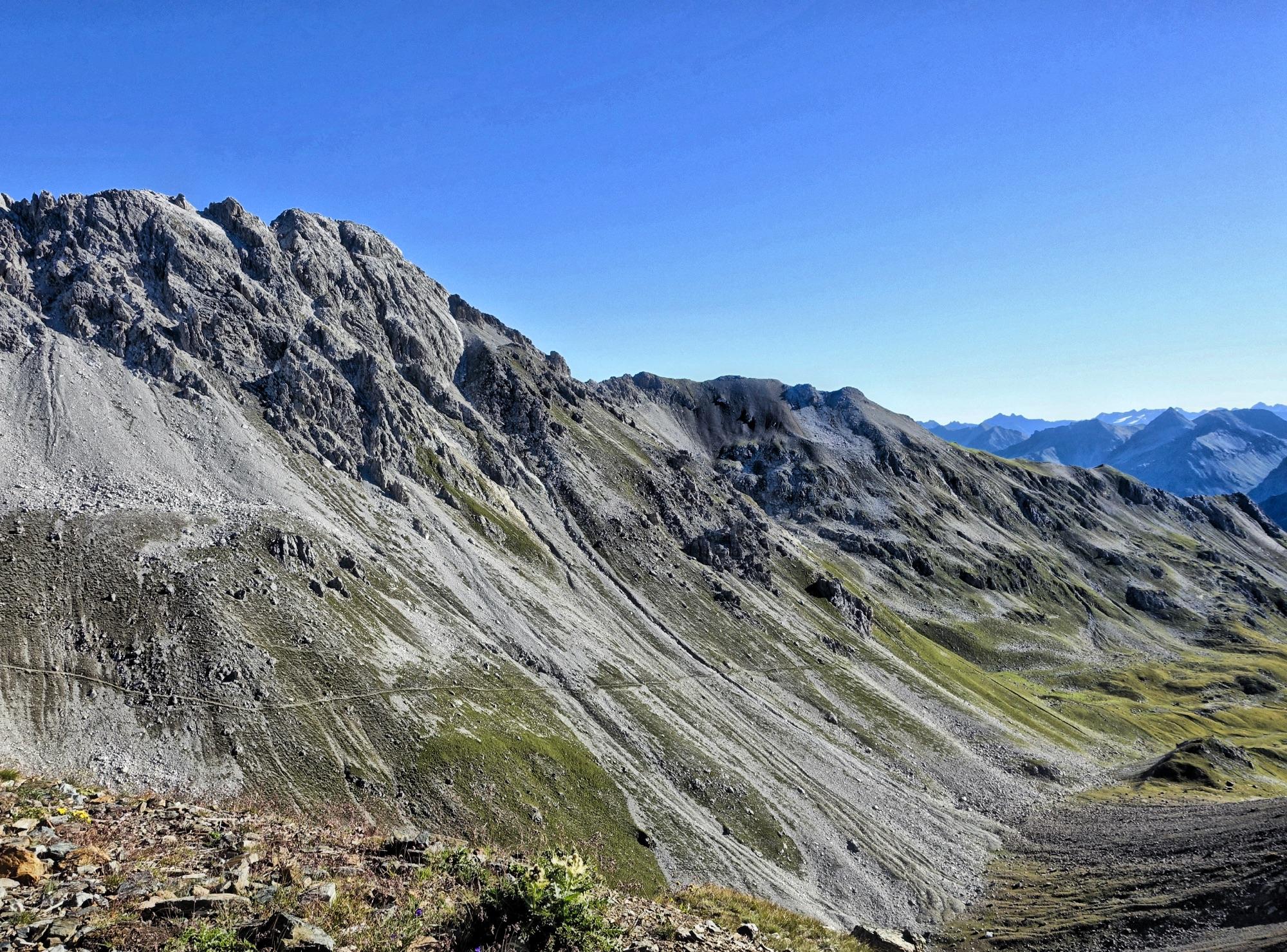 A panoramic view of rocky mountain terrain under a clear blue sky. The landscape features steep, rugged slopes with patches of green grass and rocky outcrops, showcasing the natural beauty of the mountains. Rothorn Zone mountain bike trail.