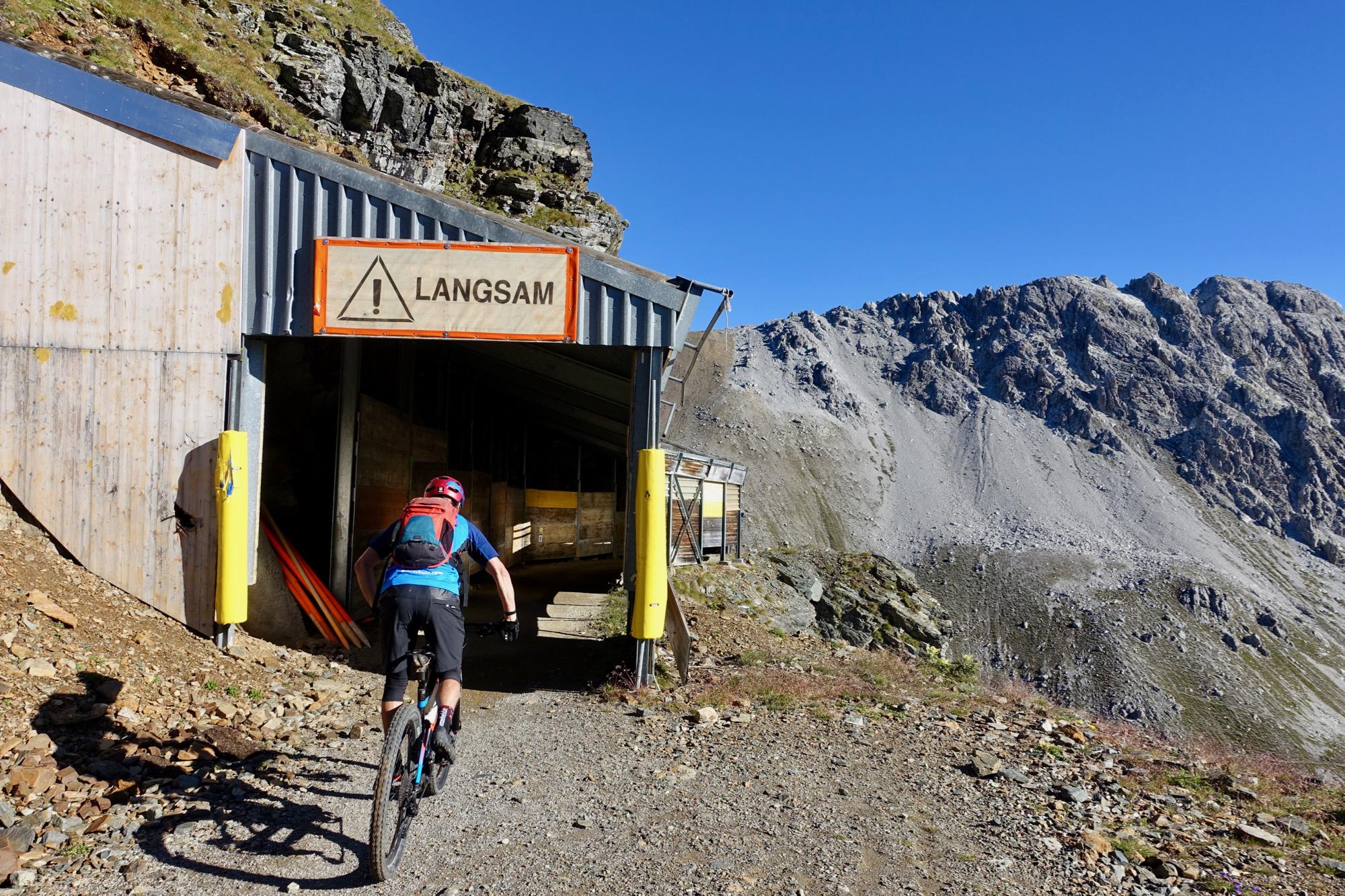 A mountain biker approaches an entryway marked with a caution sign reading "LANGSAM." The pathway leads into a dark tunnel-like structure set against a backdrop of rugged mountain terrain and a clear blue sky. The ground is covered in gravel and stones, typical of a mountainous environment. BikeTicket2Ride mountain bike trail.