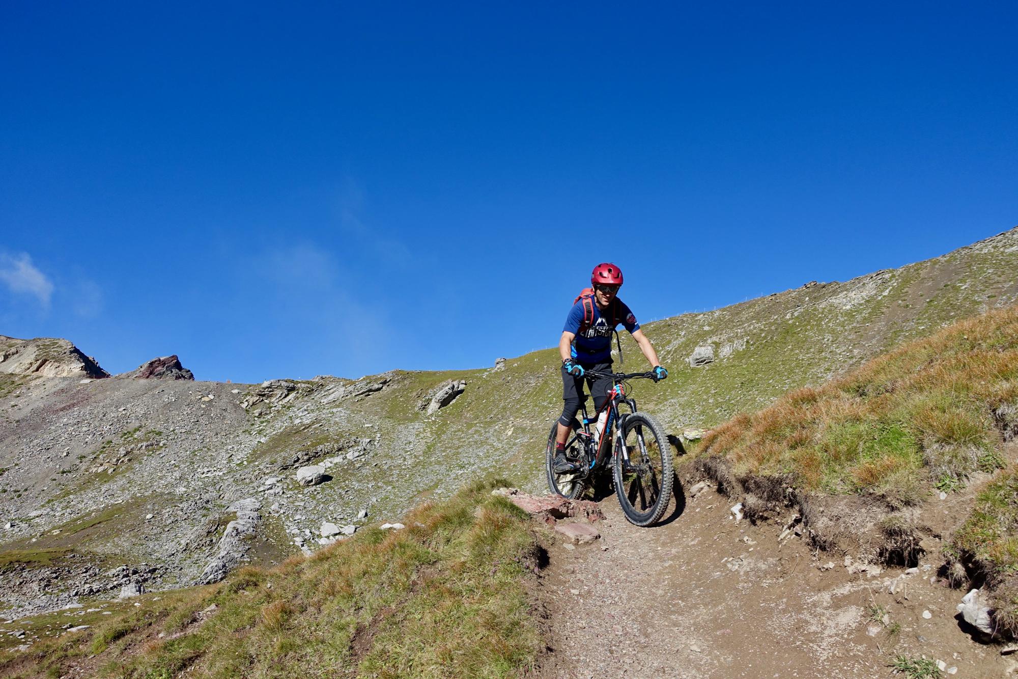 A mountain biker navigating a rocky trail on a bright sunny day, with a clear blue sky and grassy hills in the background. Rothorn Zone mountain bike trail.