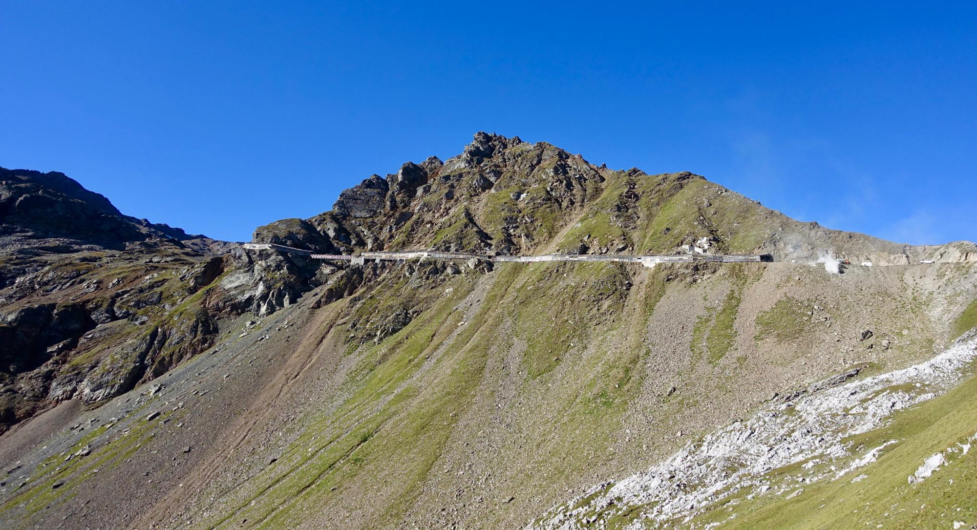 A rugged mountain landscape featuring a steep, grassy slope with rocky outcroppings. At the top, a winding road stretches along the mountainside under a clear blue sky. Rothorn Zone mountain bike trail.