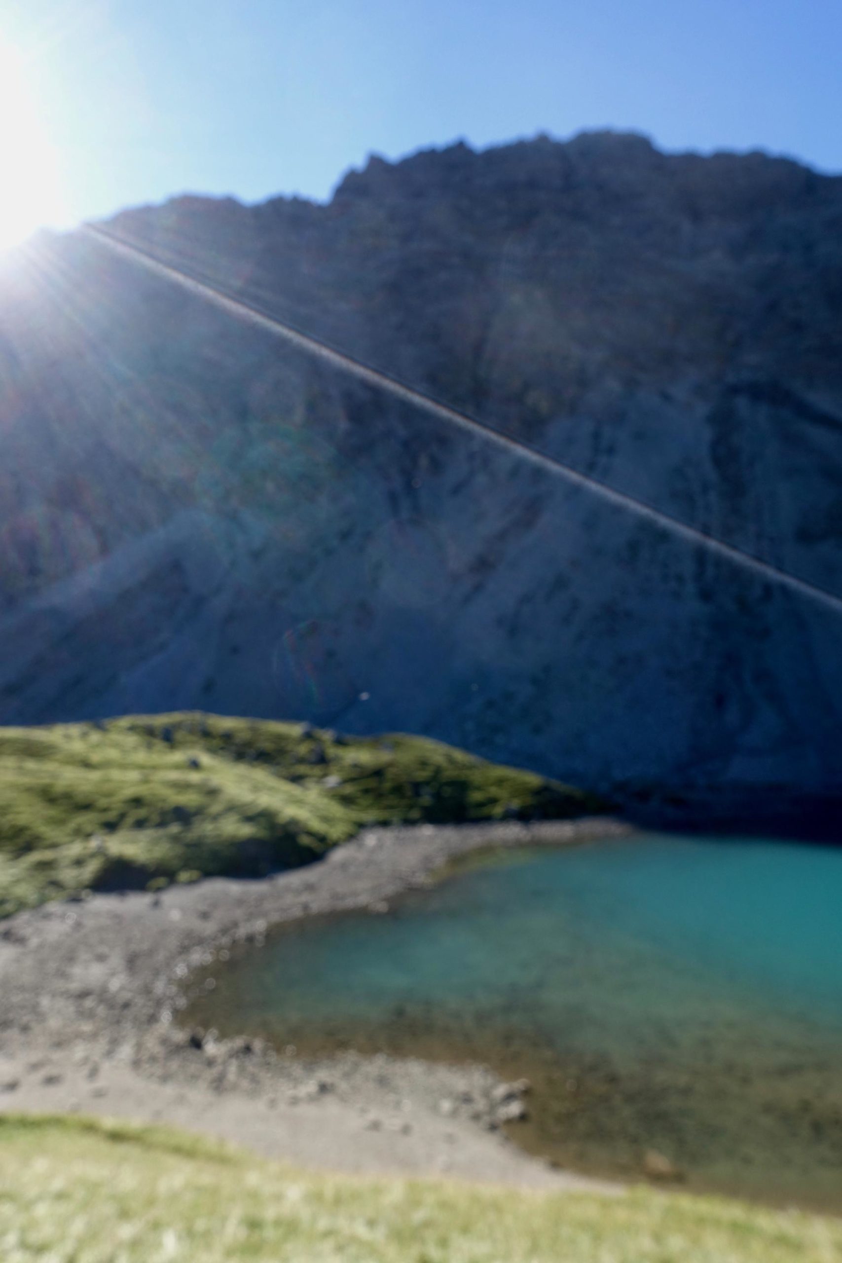 A blurred landscape photo showing a bright sun shining behind a mountain. In the foreground, there is a serene body of water with a rocky shoreline, surrounded by grassy hills. The scene captures a sense of tranquility and natural beauty. BikeTicket2Ride mountain bike trail.