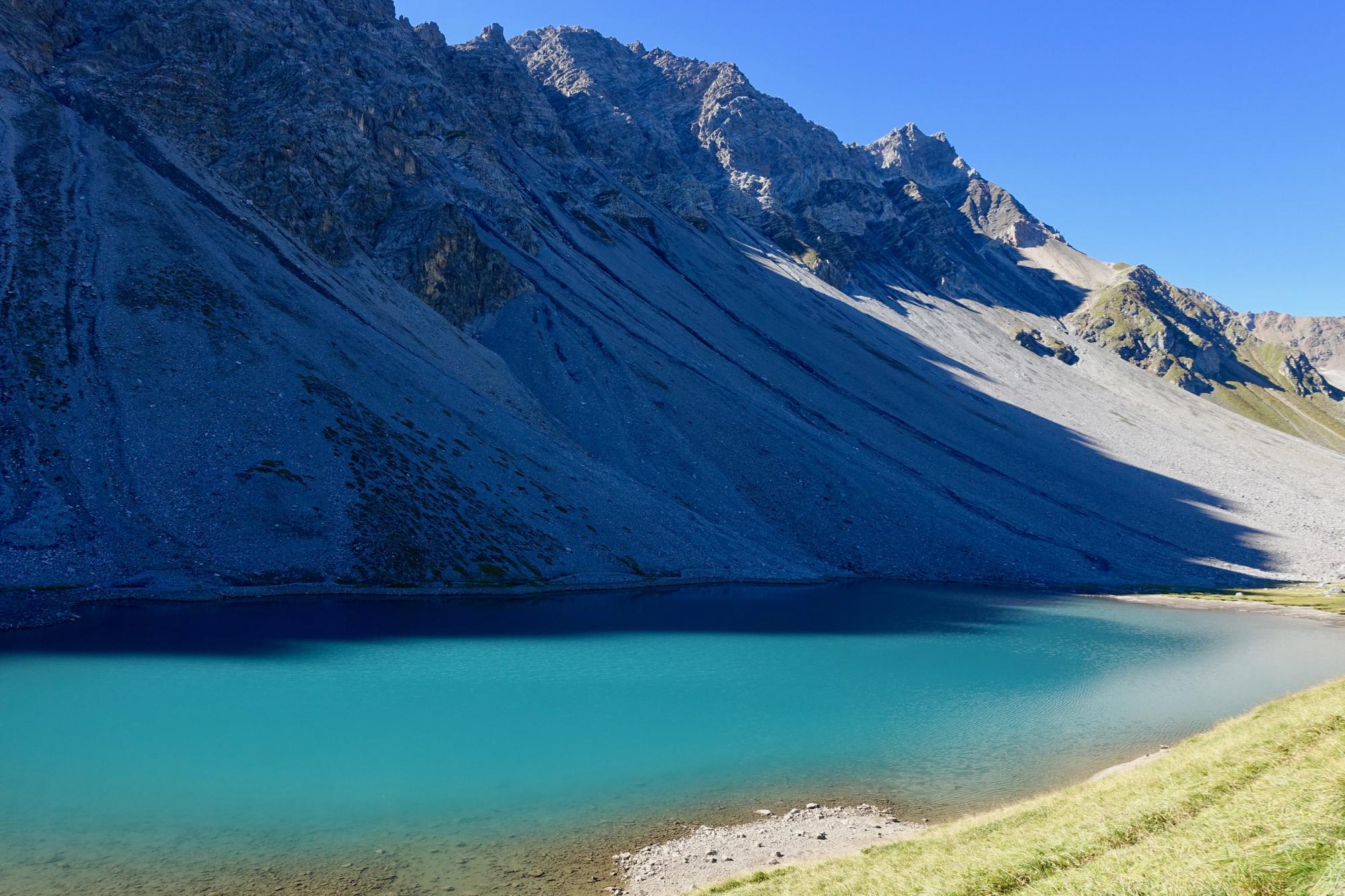 A serene mountain landscape featuring a clear, turquoise lake surrounded by steep gray rock formations and a bright blue sky. The scene showcases the natural beauty of the area, with gentle slopes leading to the water's edge and patches of green grass along the shoreline. Rothorn Zone mountain bike trail.