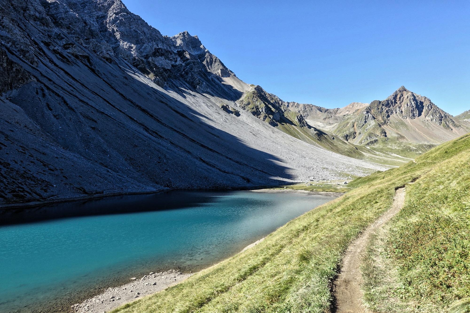 A scenic landscape featuring a clear, turquoise lake surrounded by steep, rocky mountains. The foreground includes a winding dirt path bordered by lush green grass, leading along the lake's edge. The sky is bright blue with few clouds, creating a serene atmosphere in a mountainous region. BikeTicket2Ride mountain bike trail.