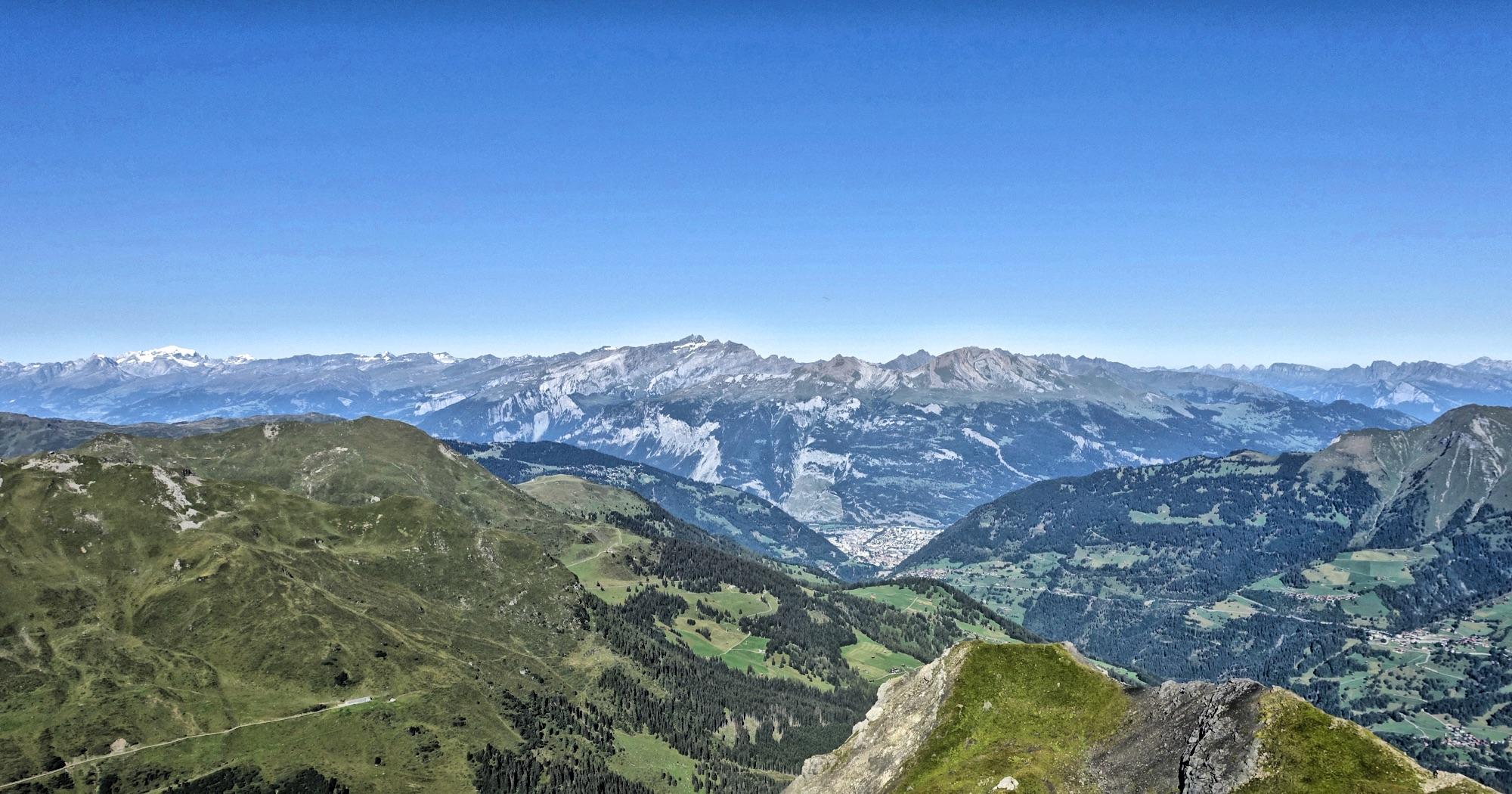 A panoramic view of majestic mountains under a clear blue sky, featuring rolling green hills and valleys in the foreground. Snow-capped peaks rise in the background, showcasing the natural beauty of the landscape. A small village is visible nestled among the lush terrain. BikeTicket2Ride mountain bike trail.