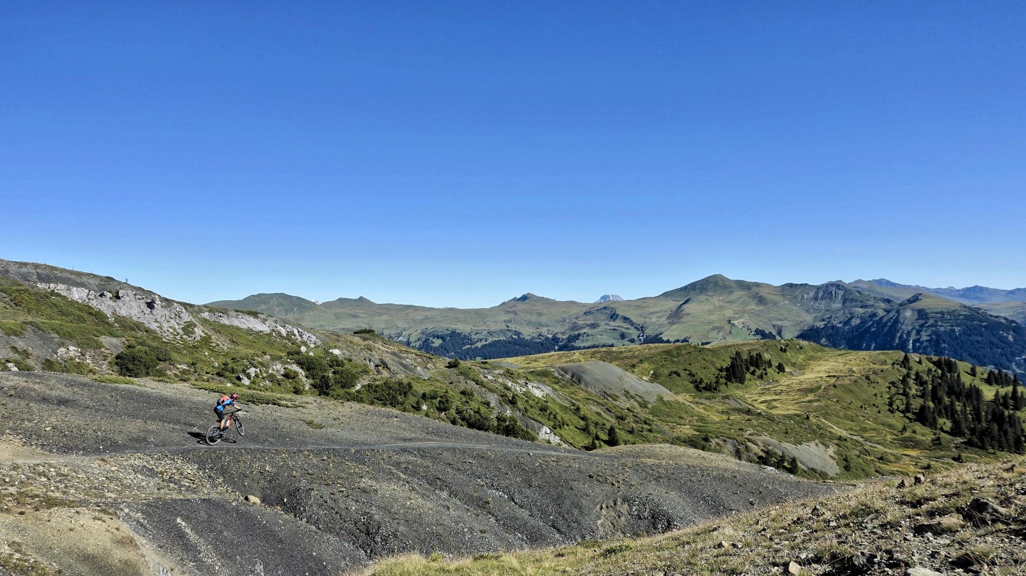 A mountain biker riding along a rugged trail in a scenic mountainous landscape under a clear blue sky. The terrain features rolling hills, rocky paths, and greenery, showcasing the beauty of outdoor adventure and nature. BikeTicket2Ride mountain bike trail.