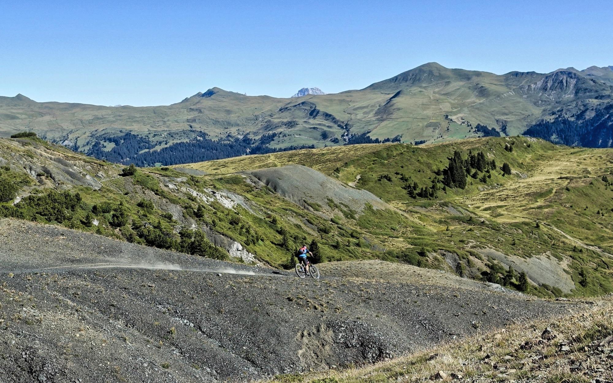 A person riding a mountain bike along a gravel path in a mountainous landscape, surrounded by rolling hills and greenery under a clear blue sky. BikeTicket2Ride mountain bike trail.