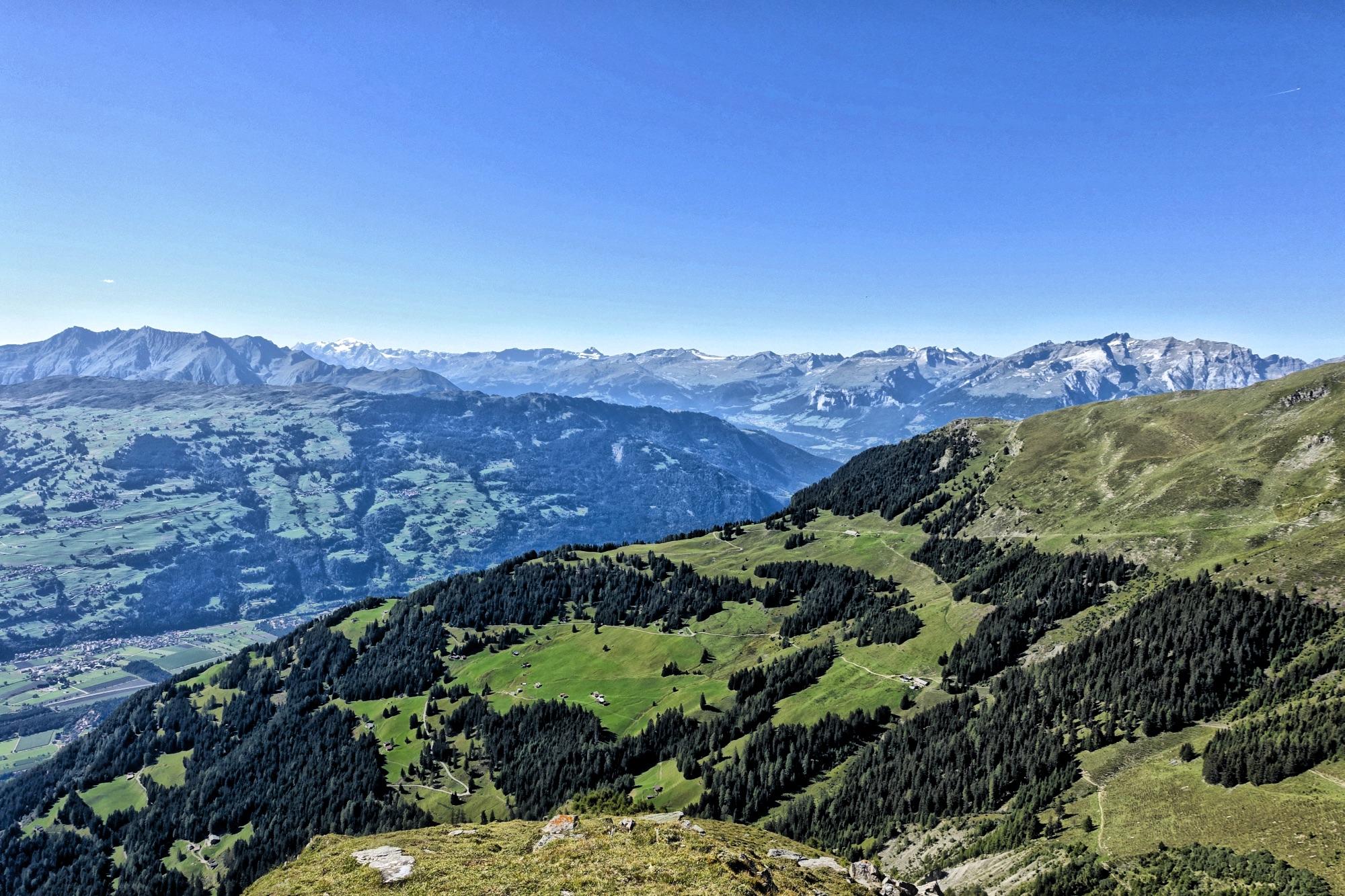A panoramic view of a mountainous landscape featuring green valleys and patches of forest, under a clear blue sky. The background showcases a range of snow-capped peaks, creating a stunning contrast with the lush greenery in the foreground. BikeTicket2Ride mountain bike trail.