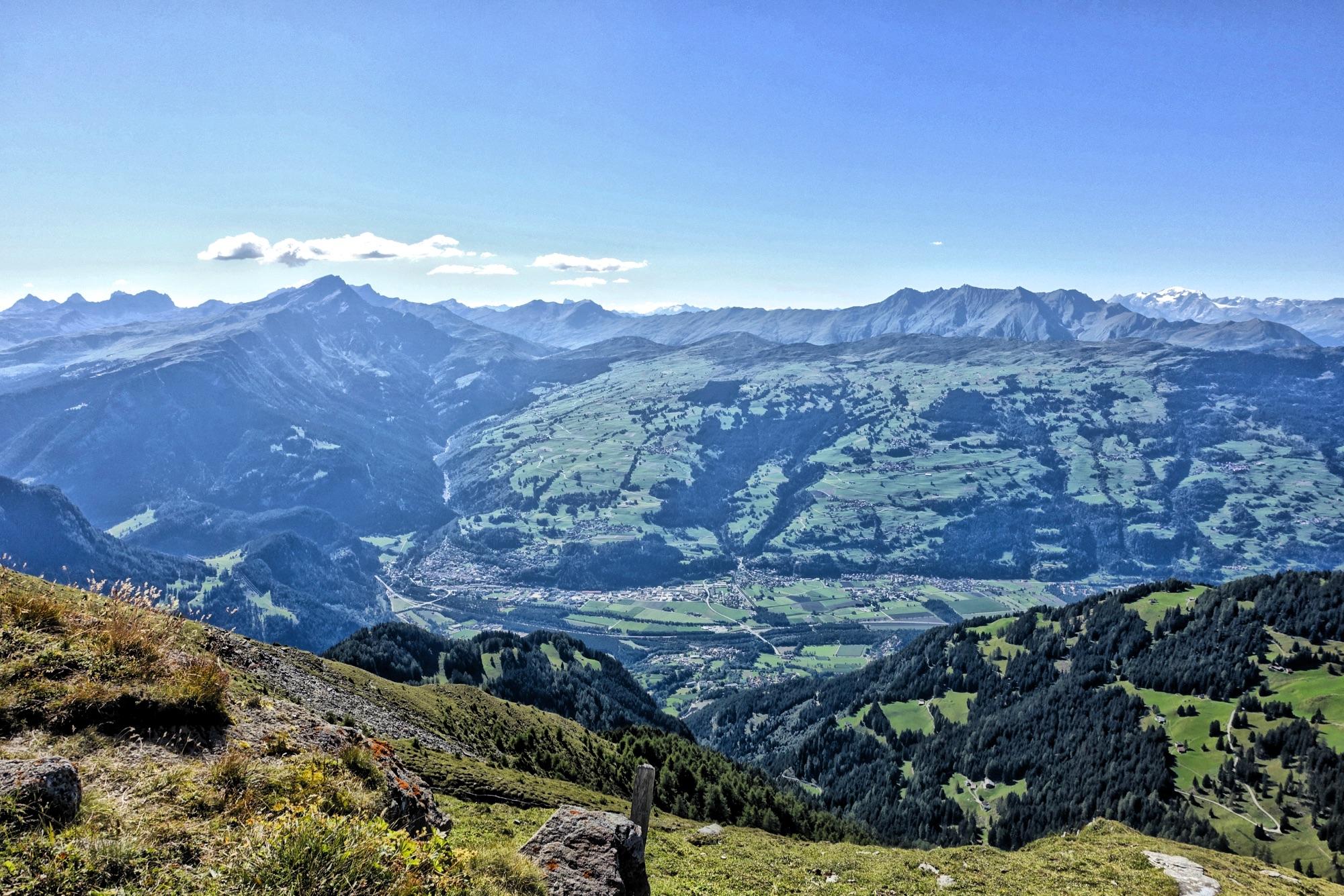 A panoramic view of a lush green valley surrounded by majestic mountains under a clear blue sky. The landscape features rolling hills, patches of forest, and winding roads, with a small town visible in the distance. Sunlight illuminates the scene, highlighting the natural beauty of the region. BikeTicket2Ride mountain bike trail.