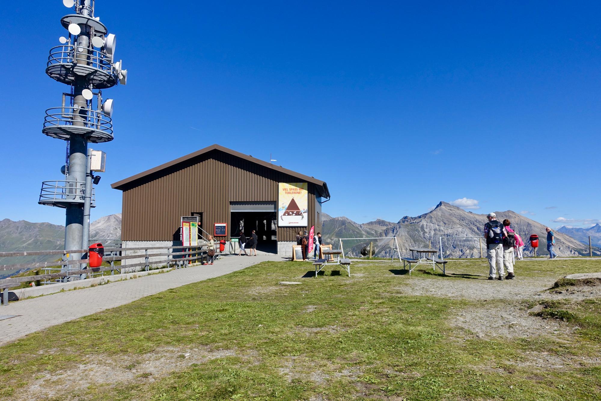 A scenic mountain view featuring a brown building with an open entrance, surrounded by green grass. A telecommunications tower stands nearby. People are walking around, with some enjoying the view of the distant mountains under a clear blue sky. Several picnic tables are set up in the foreground. BikeTicket2Ride mountain bike trail.