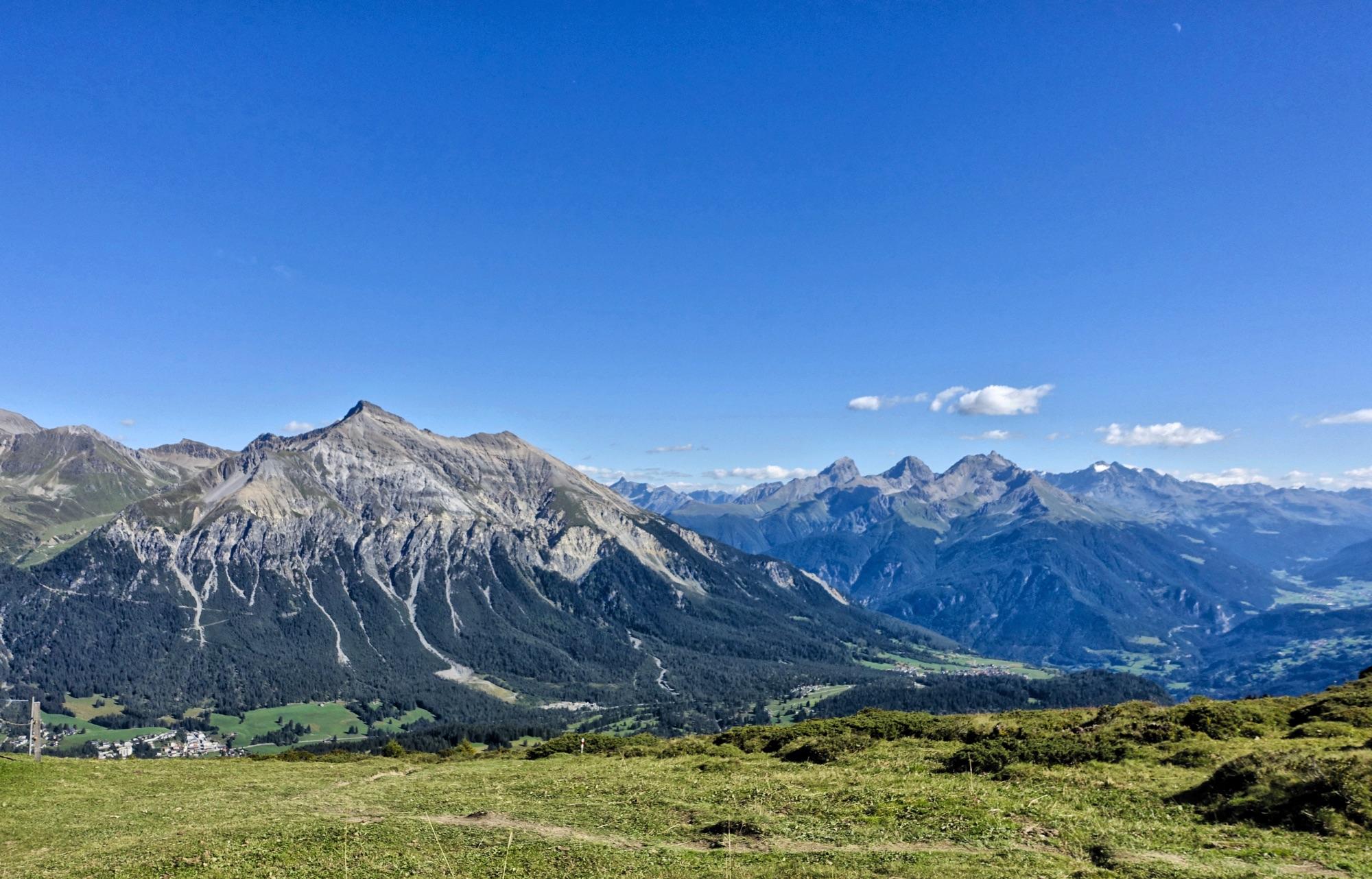 A panoramic view of majestic mountains under a clear blue sky. The landscape features rugged peaks, lush green meadows, and valleys below, showcasing the natural beauty of a mountainous region. Fluffy clouds float in the sky, adding to the serene atmosphere. BikeTicket2Ride mountain bike trail.