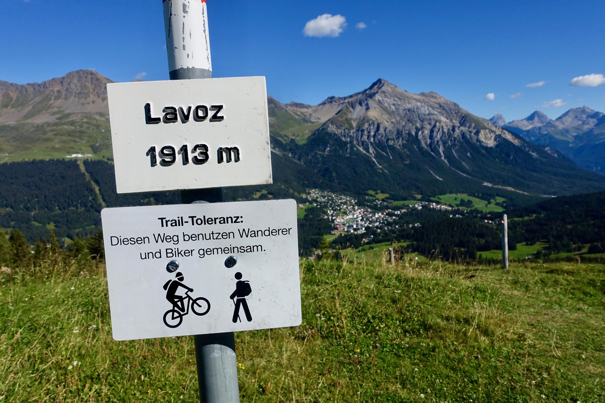 A mountain trail sign indicating "Lavoz 1913 m" with a description in German promoting shared use for hikers and bikers, set against a backdrop of mountains and a valley with a small town. BikeTicket2Ride mountain bike trail.