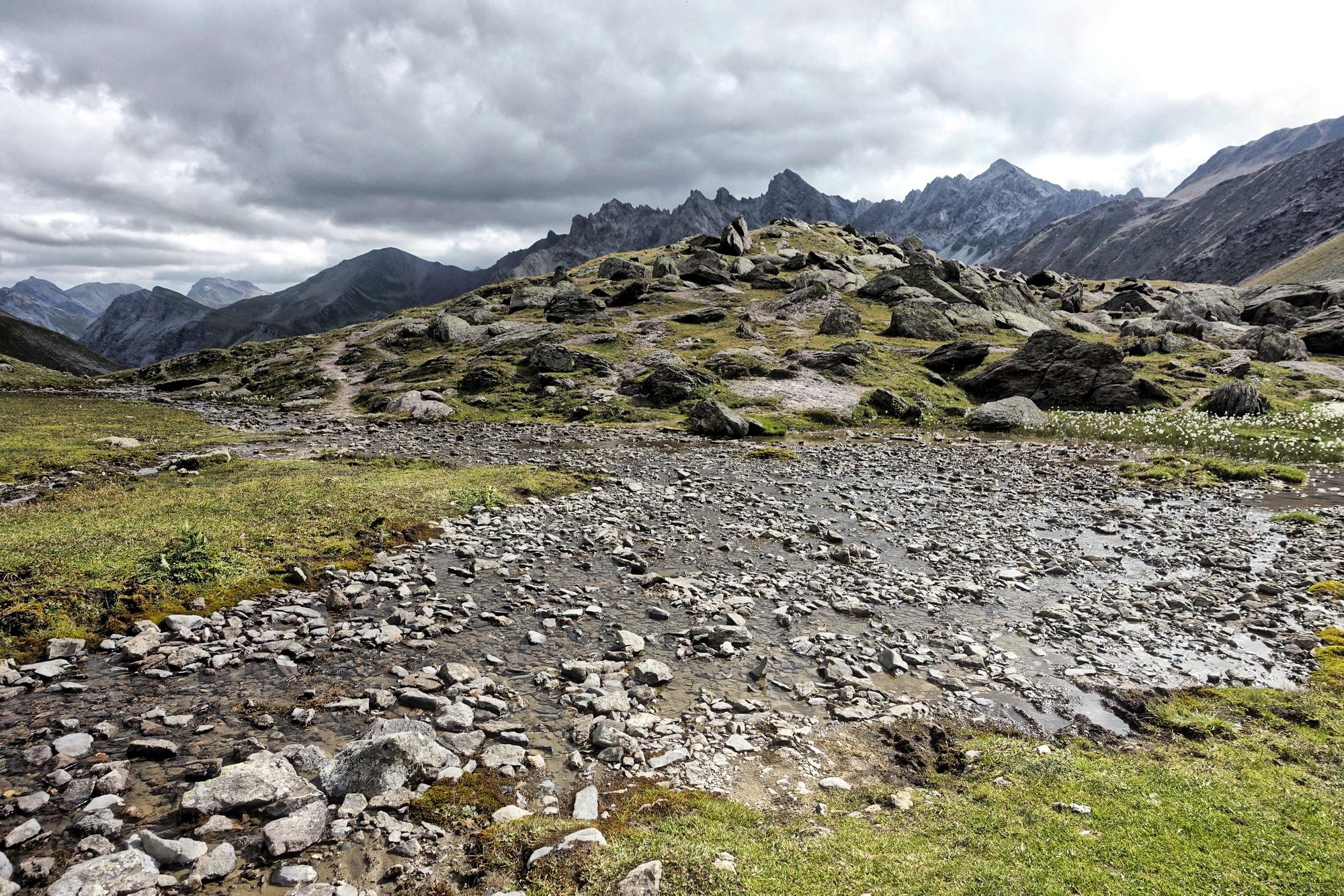 A rocky landscape featuring a winding stream and patches of grass, with a backdrop of rugged mountains under a cloudy sky. Welschtobel mountain bike trail.