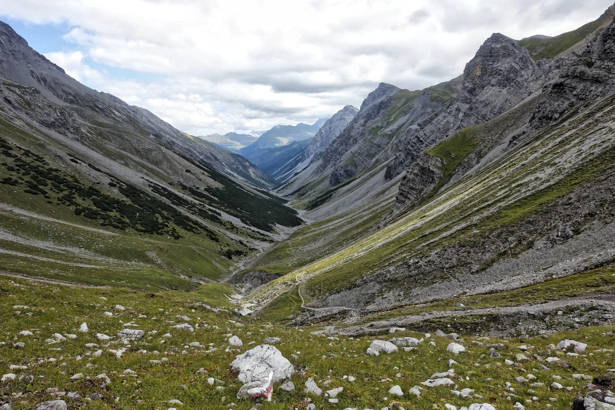 A panoramic view of a rocky valley surrounded by steep mountains, featuring green patches of vegetation and a winding path leading through the landscape. The sky is partly cloudy, enhancing the dramatic scenery of the natural environment. Welschtobel mountain bike trail.