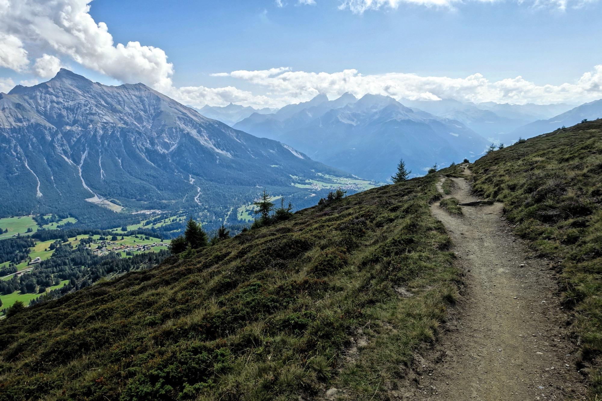 A scenic mountain landscape featuring a winding dirt path leading through grassy hills, with towering, rugged mountains in the background beneath a partly cloudy sky. Lush green valleys are visible below, showcasing a patchwork of fields and trees. BikeTicket2Ride mountain bike trail.