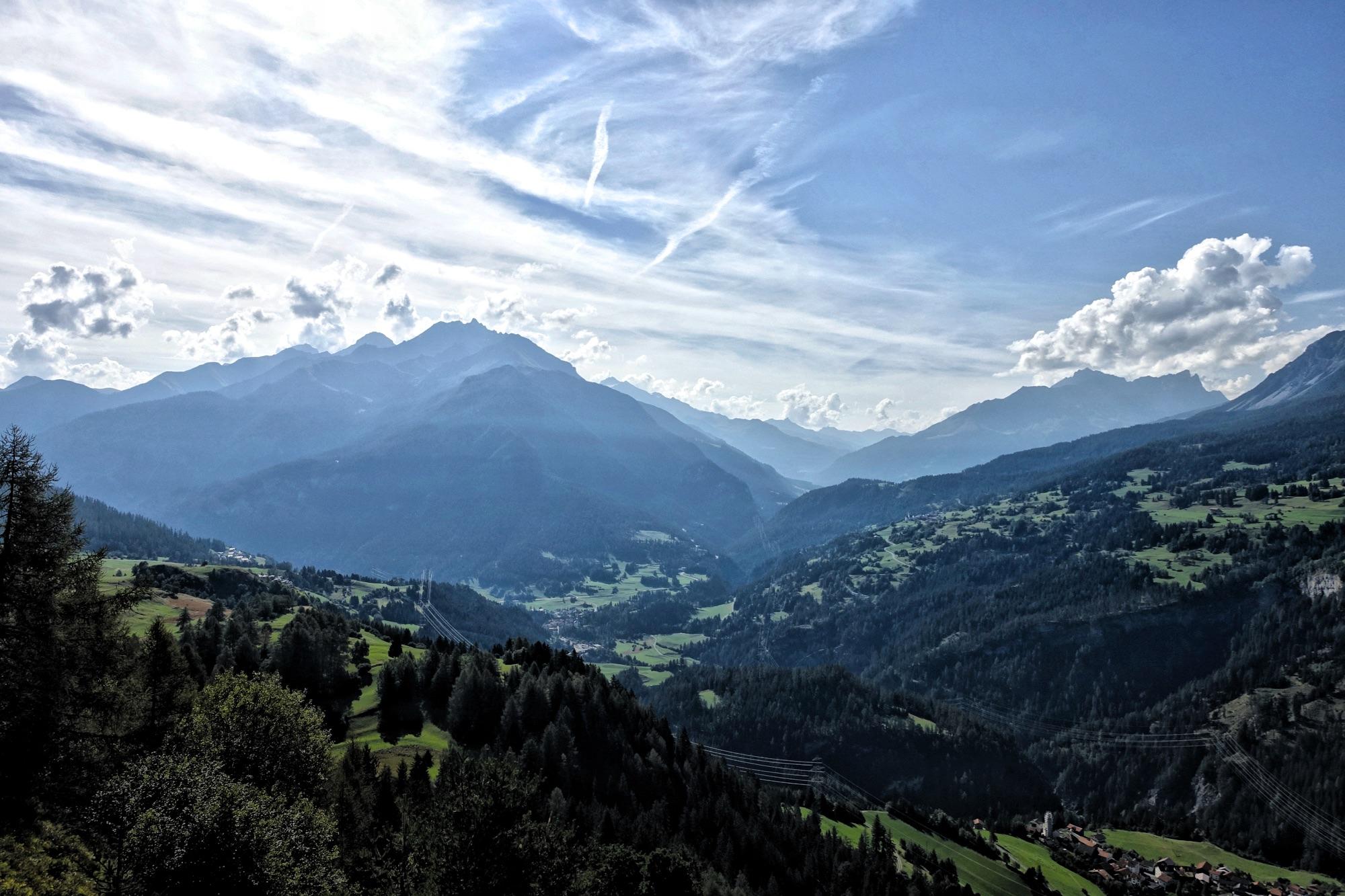 A panoramic view of a mountainous landscape, showcasing layers of peaks under a partly cloudy sky. The foreground features lush green hills and scattered trees, while a small village can be seen nestled below. The mountains in the background are shrouded in mist, creating a serene and picturesque scenery. 601 Trail mountain bike trail.