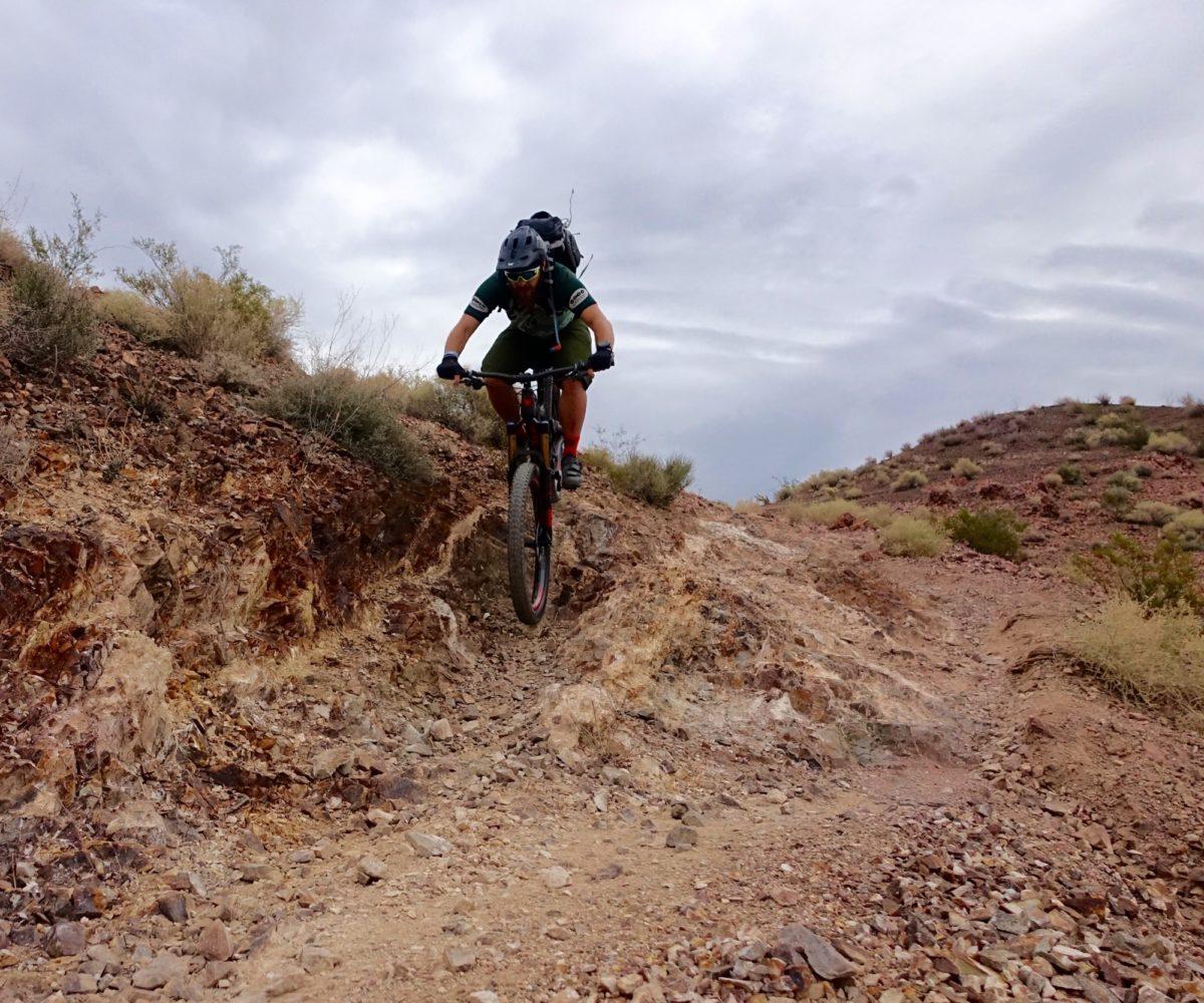 A mountain biker navigates a rocky trail, airborne as they jump over uneven terrain under a cloudy sky. The landscape features dry hills and scattered vegetation typical of a desert environment. Bootleg Canyon mountain bike trail.