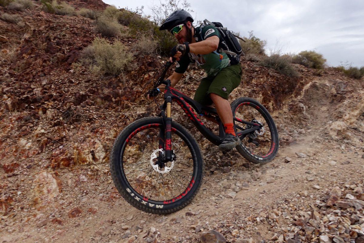 A mountain biker riding downhill on a rocky trail, surrounded by sparse vegetation and rocky terrain, with a cloudy sky in the background. The biker is wearing a helmet, sunglasses, and a backpack, showcasing an adventurous moment in a natural setting. Bootleg Canyon mountain bike trail.