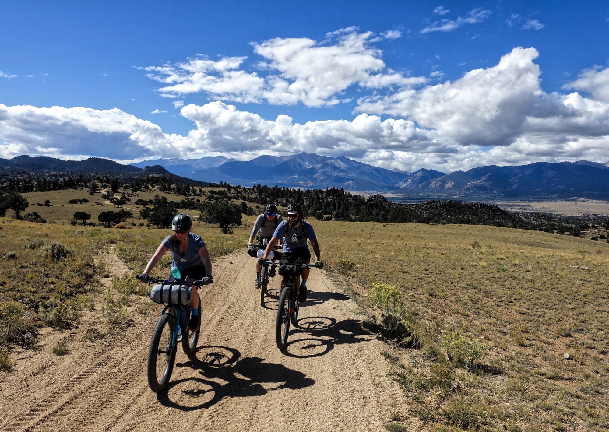 Three cyclists riding on a dirt trail through a scenic landscape featuring rolling hills and mountains in the background. The sky is partly cloudy with patches of blue. The cyclists are wearing helmets and casual outdoor clothing, and they are pedaling side by side on a sunny day. Sevenmile Creek Road / #311 mountain bike trail.
