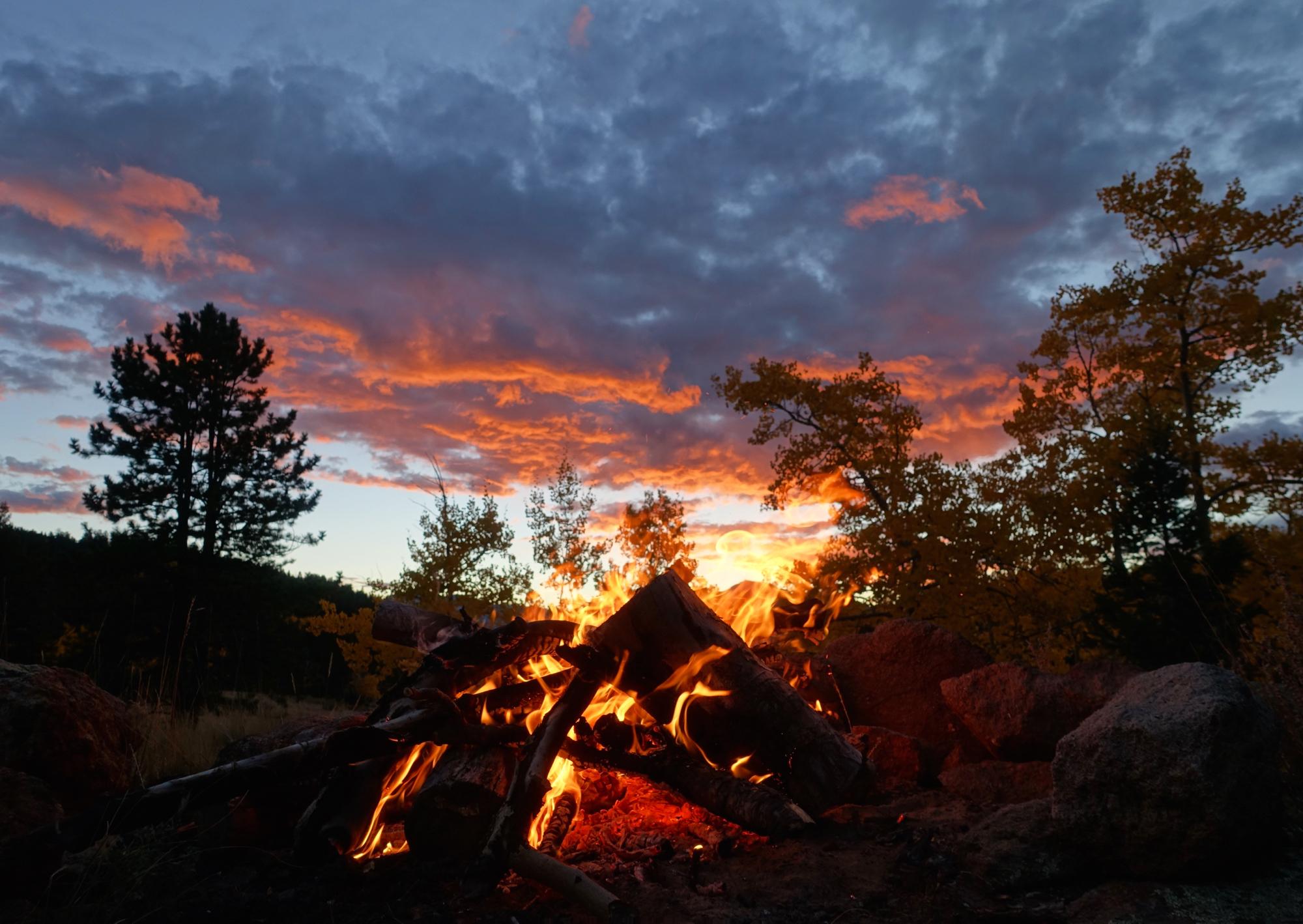 A glowing campfire surrounded by rocks, with flames and burning logs in the foreground. In the background, a vibrant sunset paints the sky with shades of orange and purple, with silhouetted pine and aspen trees. Fluffy clouds fill the sky, creating a serene and cozy outdoor atmosphere. Road #185.E mountain bike trail.
