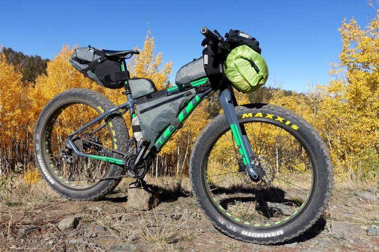 A mountain bike with green and black accents is parked on a rocky surface, surrounded by vibrant fall foliage. The bike is equipped with various bags for storage, showcasing a sturdy frame and wide tires designed for rugged terrain. Clear blue skies are visible in the background, creating a picturesque outdoor scene.