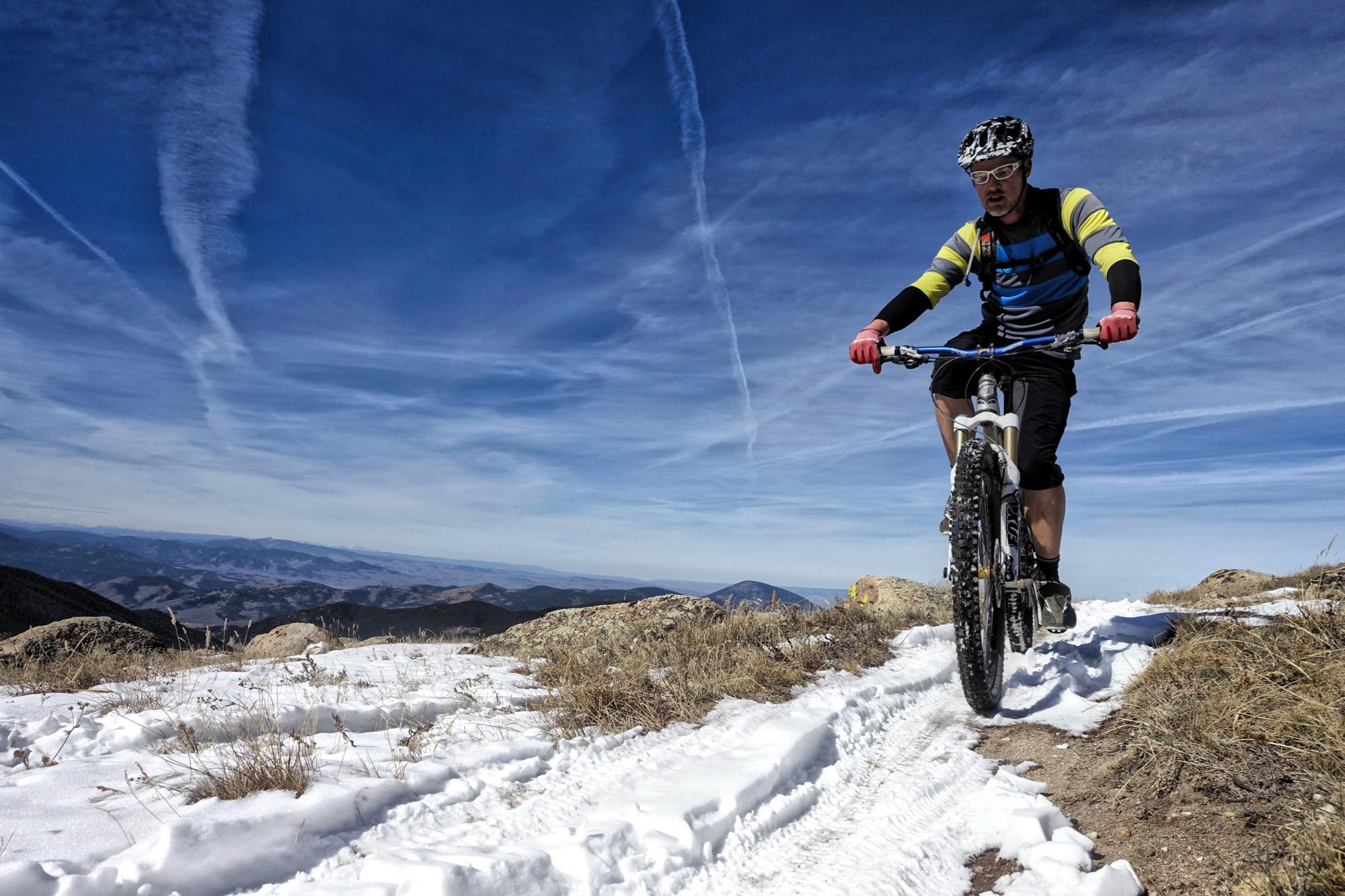 A mountain biker navigating a snowy trail on a sunny day, with a clear blue sky and distant mountains in the background. The biker is wearing a black and blue shirt with yellow stripes, gloves, and a helmet, while riding on a bike with visible tire tread marks in the snow. Monarch Crest Trail mountain bike trail.