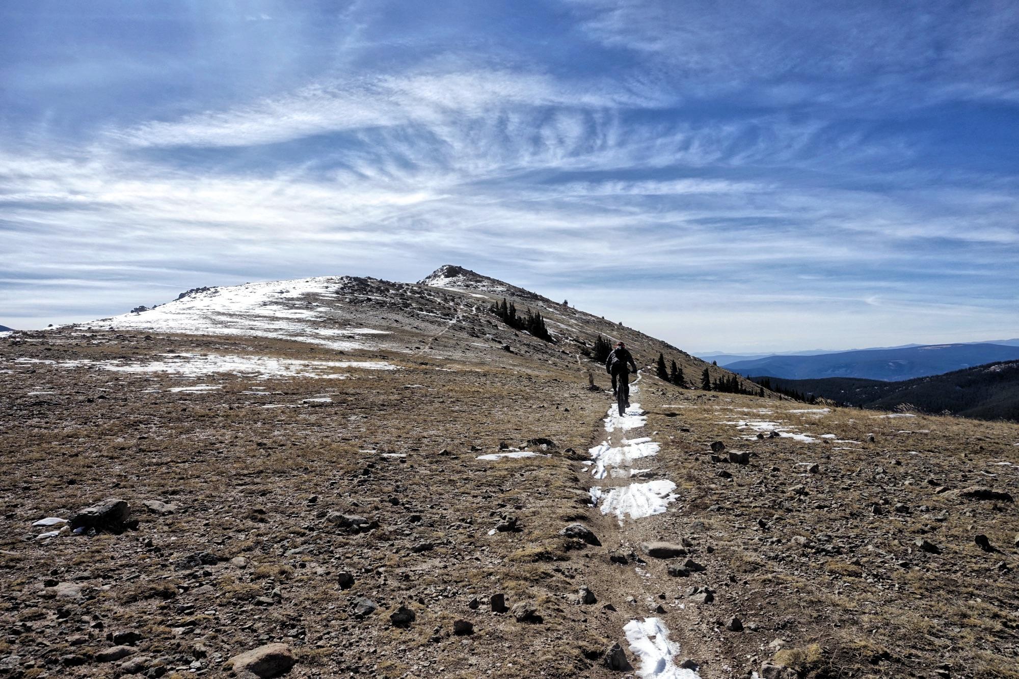 A person hiking along a rocky mountain trail with patches of snow, leading towards a snow-capped peak under a blue sky with wispy clouds. The landscape features sparse vegetation and rocky terrain, showcasing a scenic, mountainous environment. Monarch Crest Trail mountain bike trail.