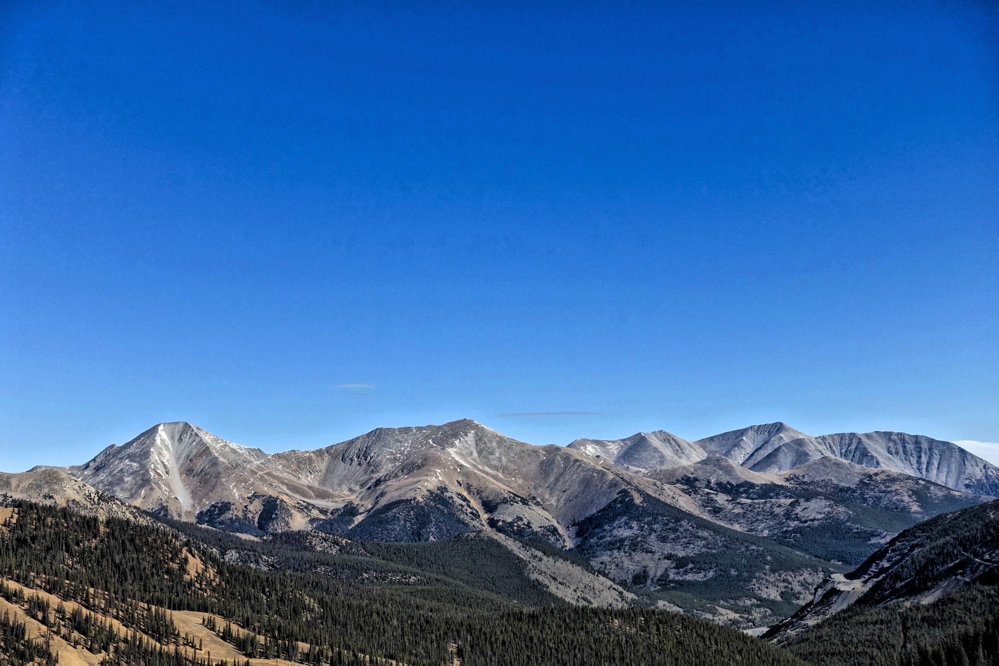 A panoramic view of a mountain range under a clear blue sky, featuring rugged peaks and sloping hills covered with dense forests. The landscape showcases a mix of rocky surfaces and patches of snow on higher elevations. CDT: Monarch Pass to Boss Lake / Hunt Lake Trail mountain bike trail.