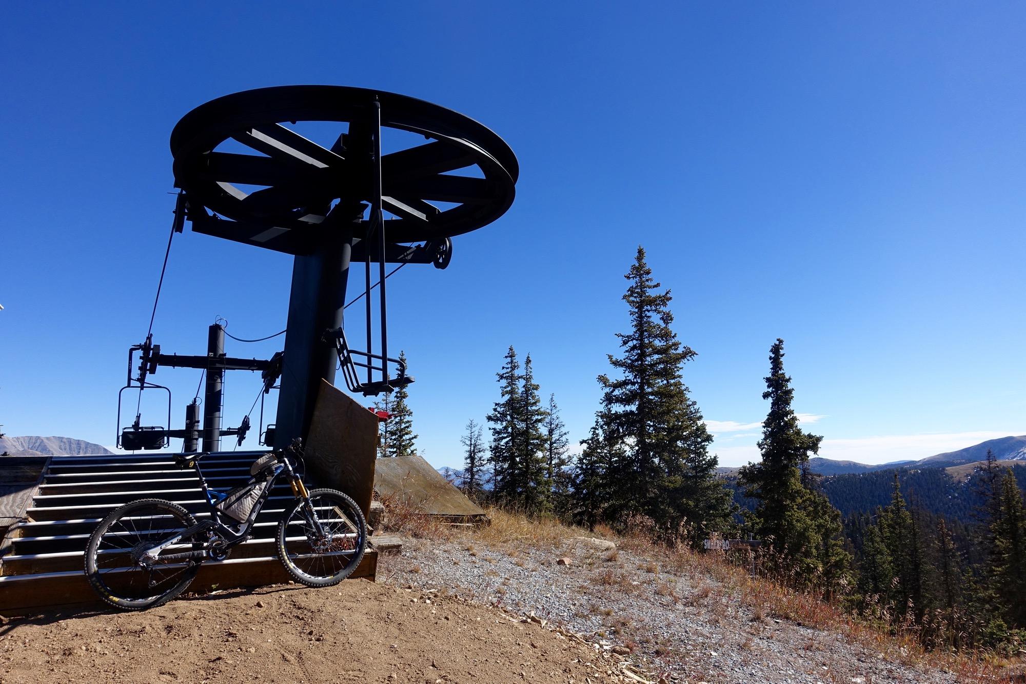 A mountain bike resting on the ground next to a ski lift at a high-altitude location, surrounded by pine trees and under a clear blue sky. CDT: Monarch Pass to Boss Lake / Hunt Lake Trail mountain bike trail.