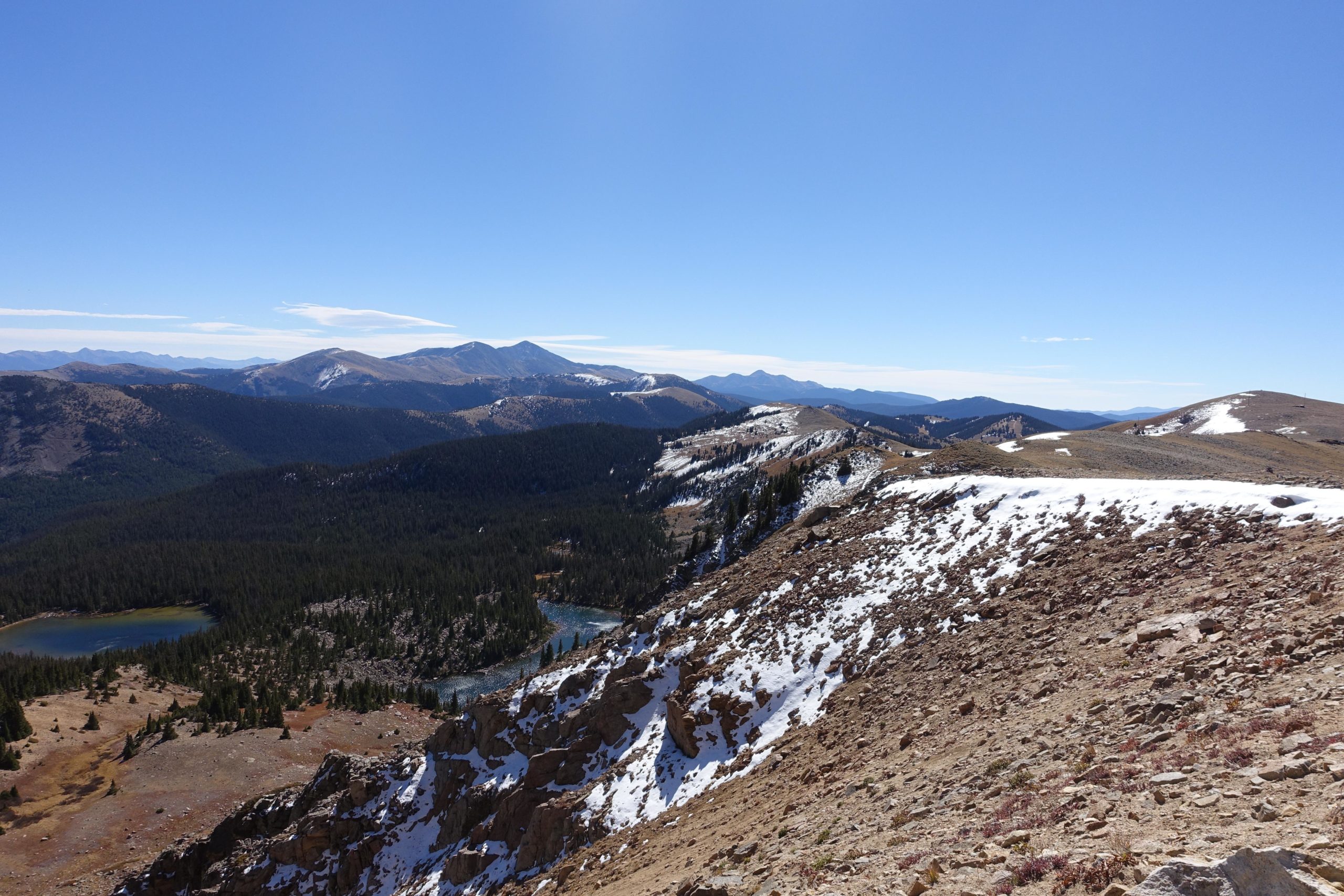 A panoramic view of mountainous terrain under a clear blue sky. The foreground features rocky, snow-dusted hills, while lush green forests and a winding river are visible in the valley below. In the background, more mountains rise sharply, creating a stunning backdrop to the serene landscape. CDT: Monarch Pass to Boss Lake / Hunt Lake Trail mountain bike trail.