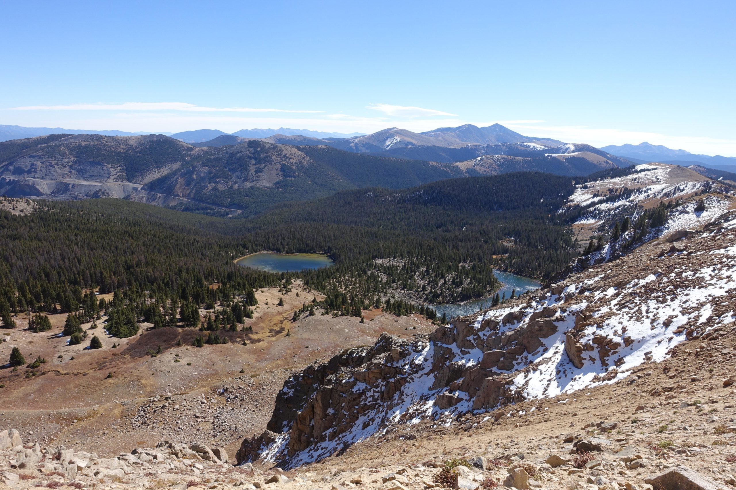 A panoramic view of a mountainous landscape featuring two lakes surrounded by dense coniferous forests, with rocky terrain and patches of snow on the ground. The clear blue sky provides a contrast to the earthy tones of the land below. The backdrop consists of rolling mountains, creating a picturesque natural scene. CDT: Monarch Pass to Boss Lake / Hunt Lake Trail mountain bike trail.