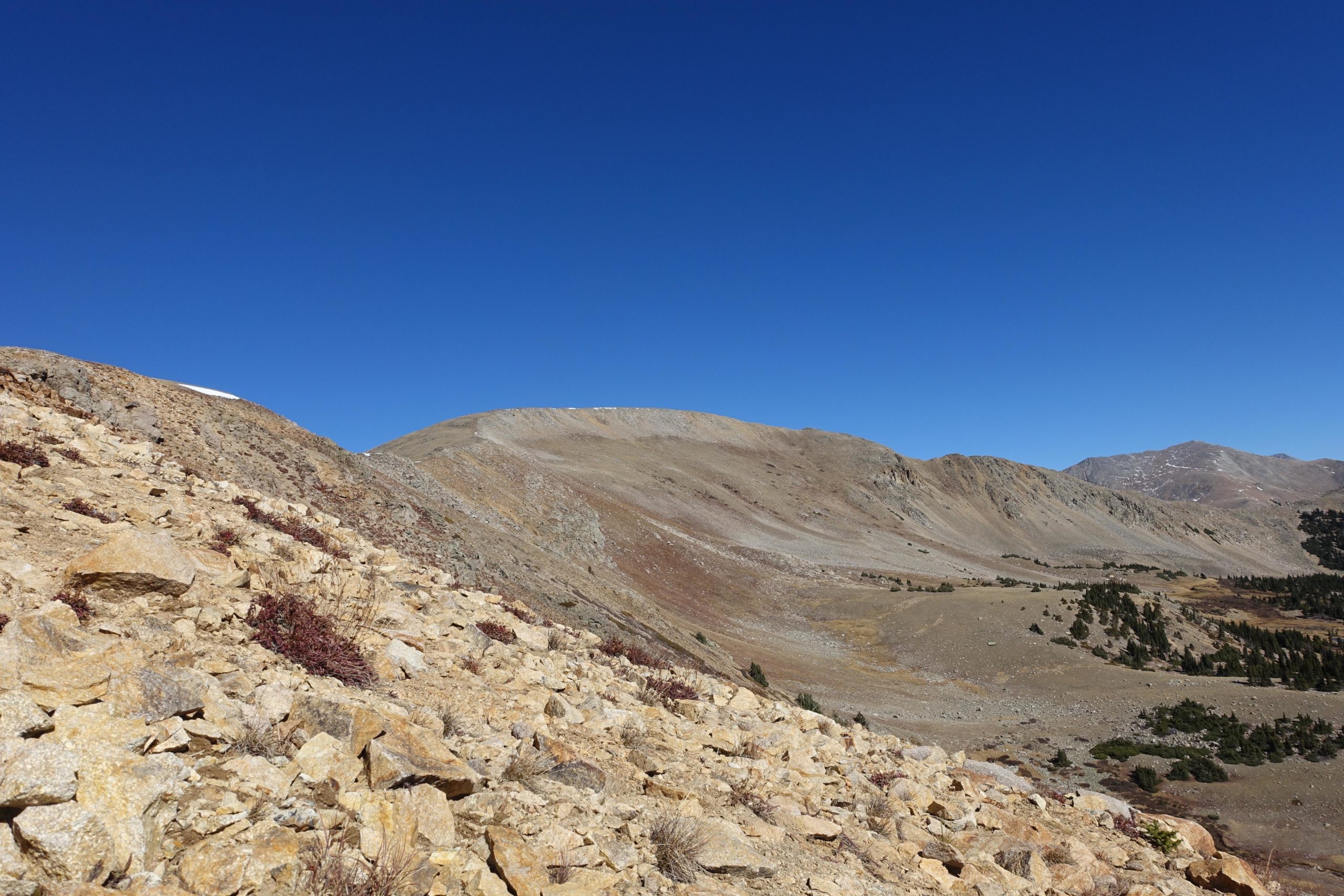 A rocky hillside leading to distant mountains under a clear blue sky, with patches of greenery in the valley below. CDT: Monarch Pass to Boss Lake / Hunt Lake Trail mountain bike trail.