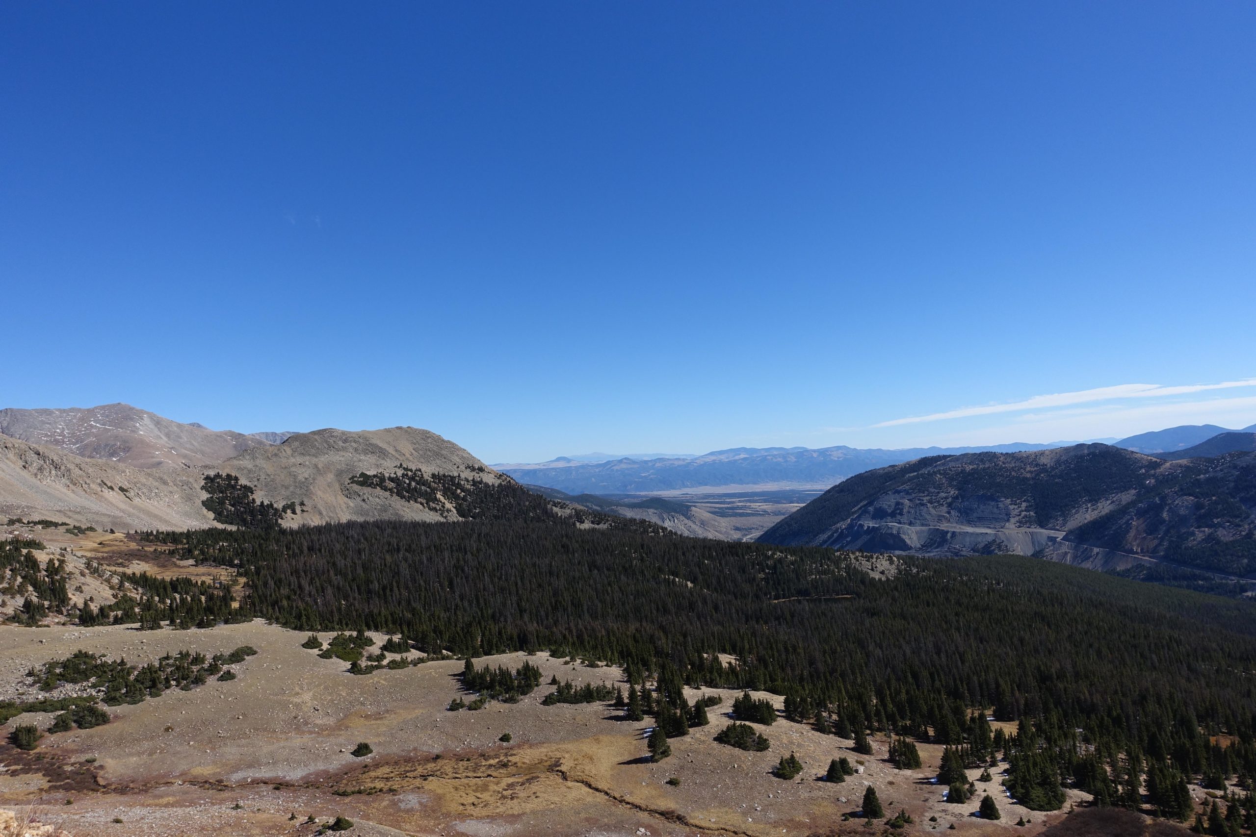 A panoramic view of a mountainous landscape, featuring rolling hills and rocky terrain with scattered evergreen trees. A clear blue sky stretches overhead, creating a serene backdrop for the distant mountain range. The foreground shows a mix of brown and green hues, highlighting the natural beauty of the wilderness area. CDT: Monarch Pass to Boss Lake / Hunt Lake Trail mountain bike trail.