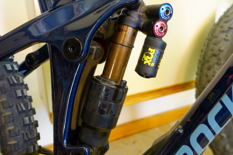 Close-up view of a bicycle's rear suspension system, featuring a Fox Float shock absorber integrated into the frame. The image showcases the shock's adjustable settings and mounting points, with visible dirt on the tire and frame, indicating usage in off-road conditions.