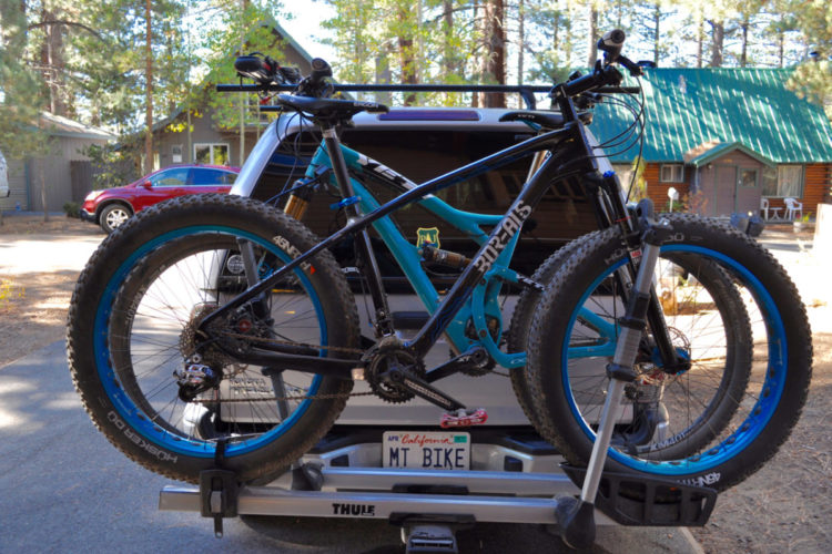 A black and blue fat tire mountain bike mounted on a bike rack attached to the back of a vehicle, with a cabin in the background. The bike features wide tires and a robust frame, showcasing the details of the drivetrain and suspension. The setting appears to be in a wooded area, with trees and other vehicles visible nearby.