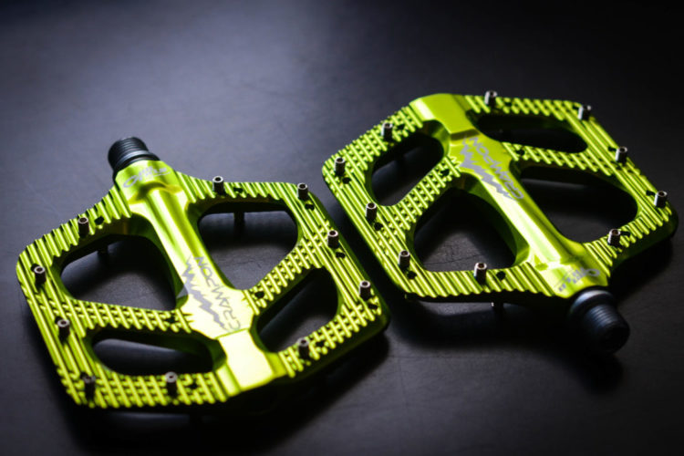 Bright green bicycle pedals with a textured surface and multiple grip pins, positioned on a dark background.