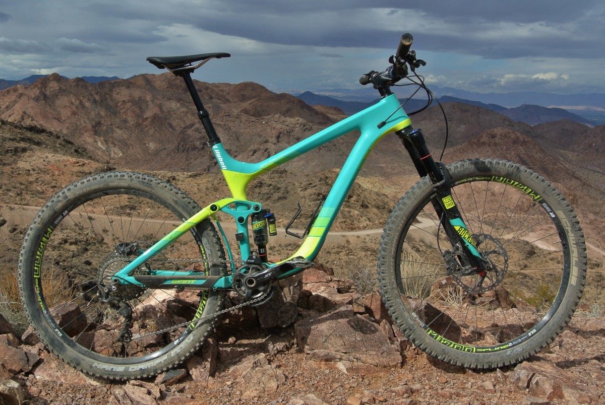 A brightly colored mountain bike resting on rocky terrain with a mountainous landscape in the background. The bike features a turquoise and lime green frame, wide tires with knobby tread, and is positioned at an angle to showcase its design and surroundings.