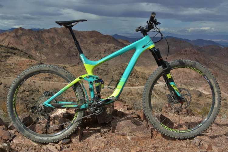 A brightly colored mountain bike resting on rocky terrain with a mountainous landscape in the background. The bike features a turquoise and lime green frame, wide tires with knobby tread, and is positioned at an angle to showcase its design and surroundings.