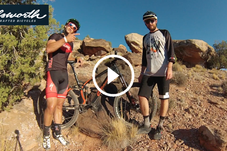 Two mountain bikers posing next to their bikes in a rocky outdoor setting. One biker is giving a thumbs-up gesture, while the other stands beside him, both wearing cycling gear. The background includes shrubs and rocky terrain, with a clear blue sky above. The Ellsworth logo for handcrafted bicycles is displayed at the top of the image.