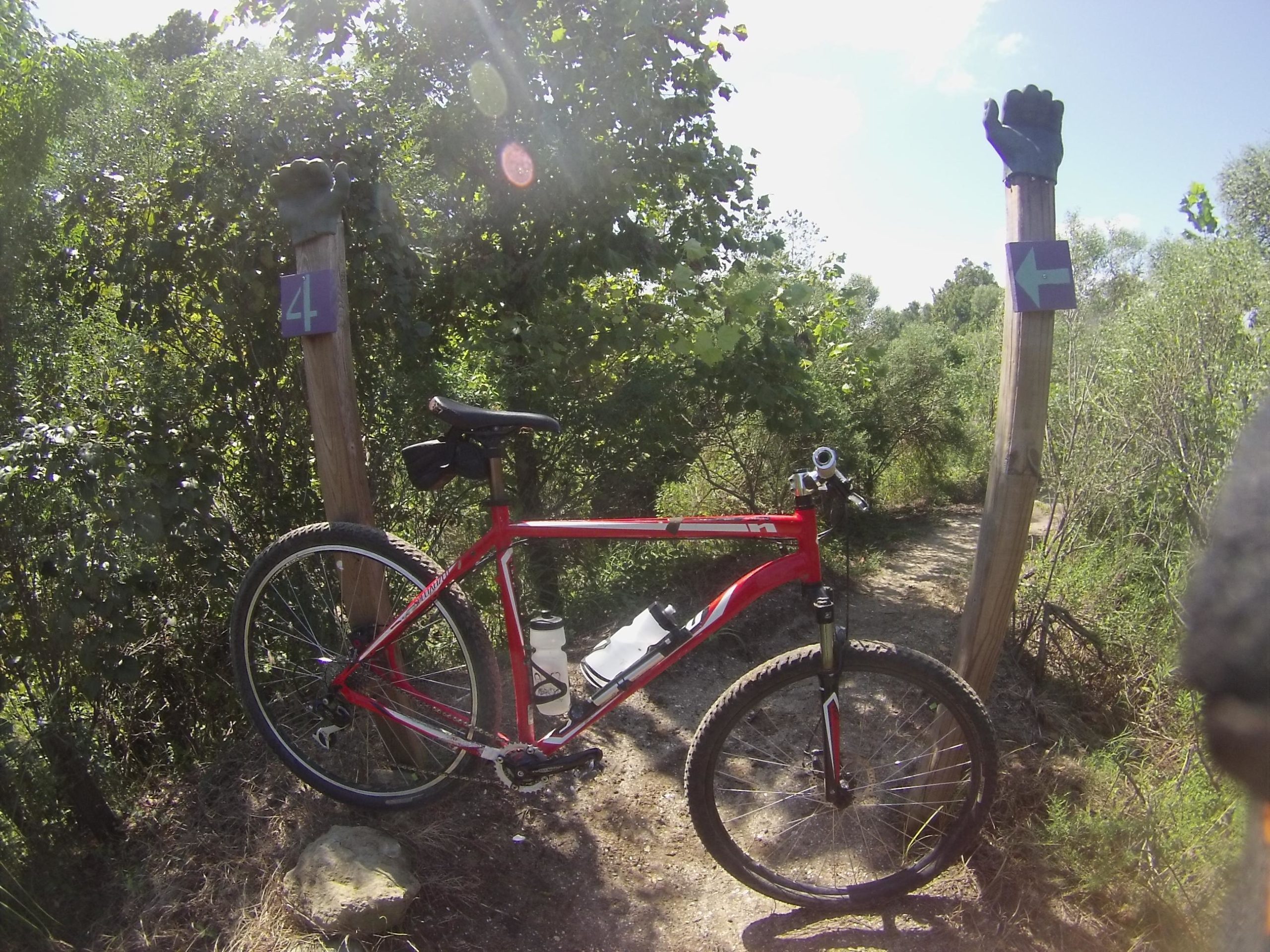 A red mountain bike is parked beside a wooden signpost on a dirt trail, surrounded by dense greenery. The sign features the number "4" and a left-pointing arrow, with a raised black hand symbolizing direction. Sunlight filters through the trees, creating a bright and inviting outdoor atmosphere. Horry County Bike Run Park mountain bike trail.
