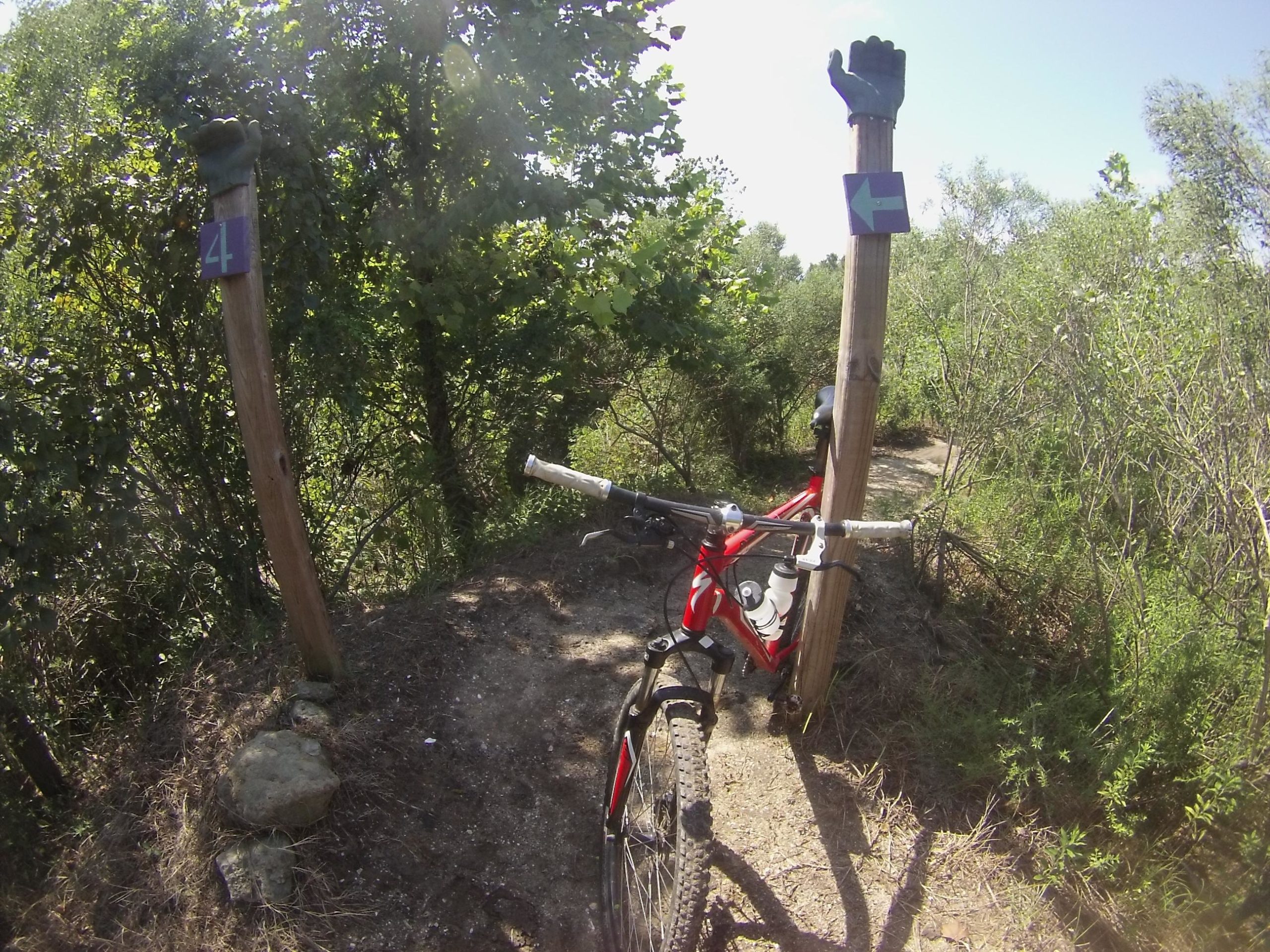 Image of a mountain bike parked on a dirt path surrounded by greenery. Two wooden posts are visible, one displaying the number "4" and the other featuring a sign with an arrow pointing left. The scene captures a sunny day in a biking area, emphasizing the trail's natural surroundings. Horry County Bike Run Park mountain bike trail.