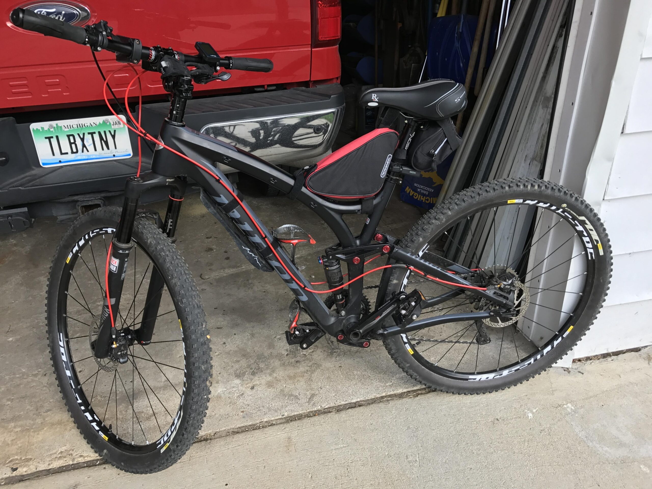 Niner Jet 9 Carbon: A mountain bike with a black and red frame, featuring thick knobby tires and front suspension, is parked next to a red vehicle in a garage setting. The bike has a small bag attached to the seat and is positioned against the garage wall, with various tools and equipment visible in the background.