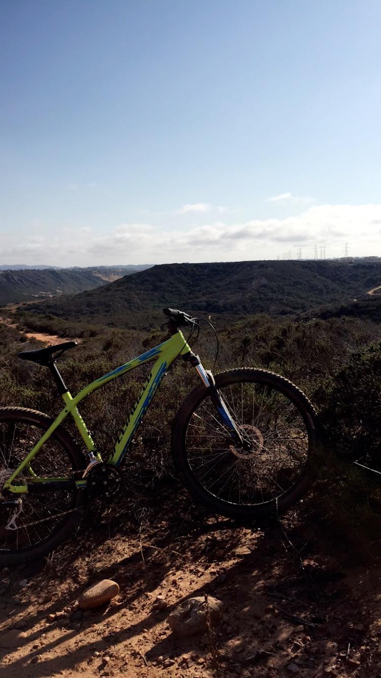 Specialized Pitch Sport 650B: A mountain bike resting on a rocky trail overlooking a scenic valley, with rolling hills in the background and power lines visible in the distance under a clear blue sky.