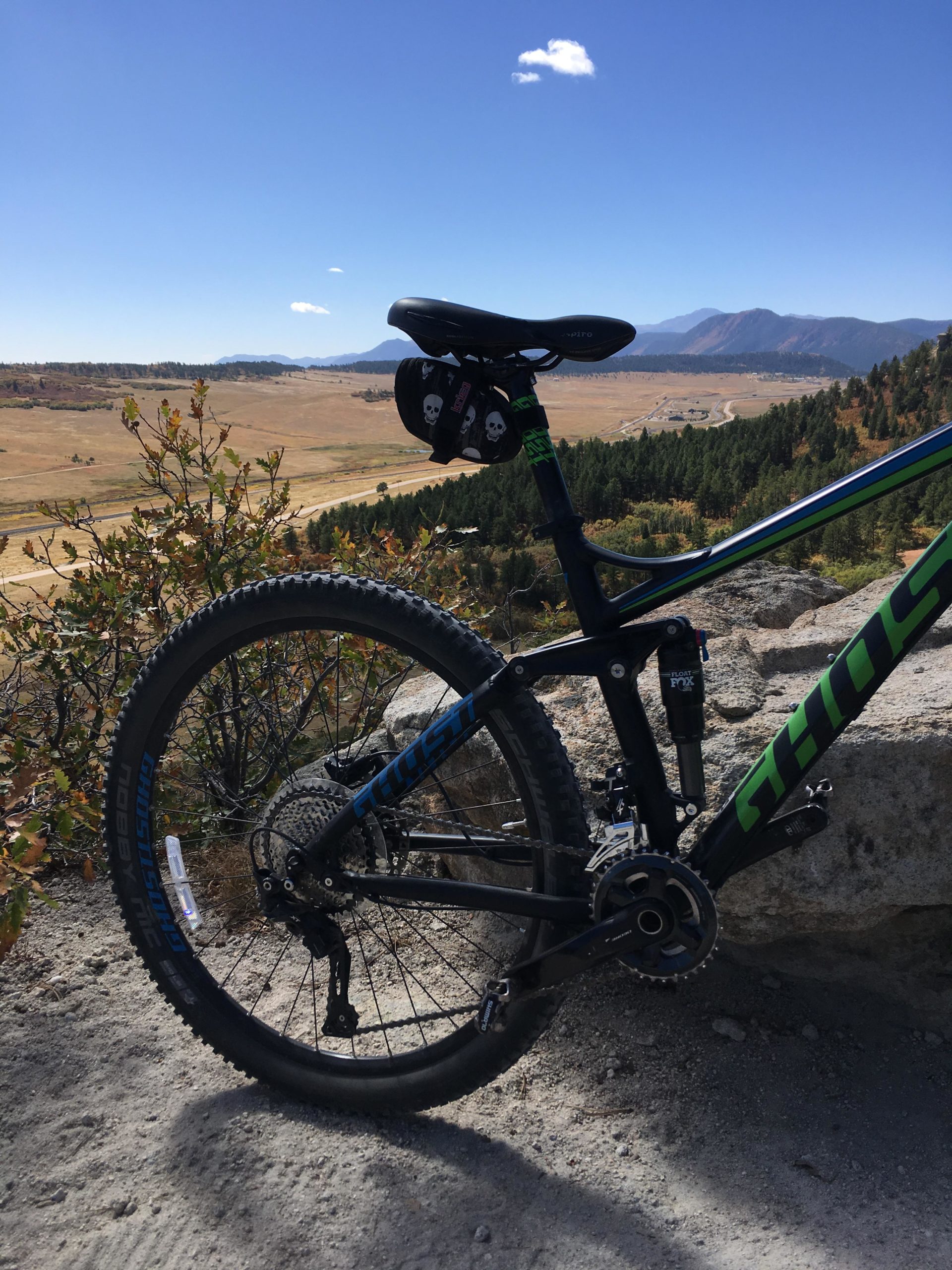 A close-up view of a mountain bike seated on a rocky surface, with a scenic backdrop of rolling hills and a clear blue sky. The bike features a black and green frame with visible branding, and the seat has headphones hanging from it. In the distance, mountains rise under a few wispy clouds. Spruce Mountain Trail Upper Loop mountain bike trail.