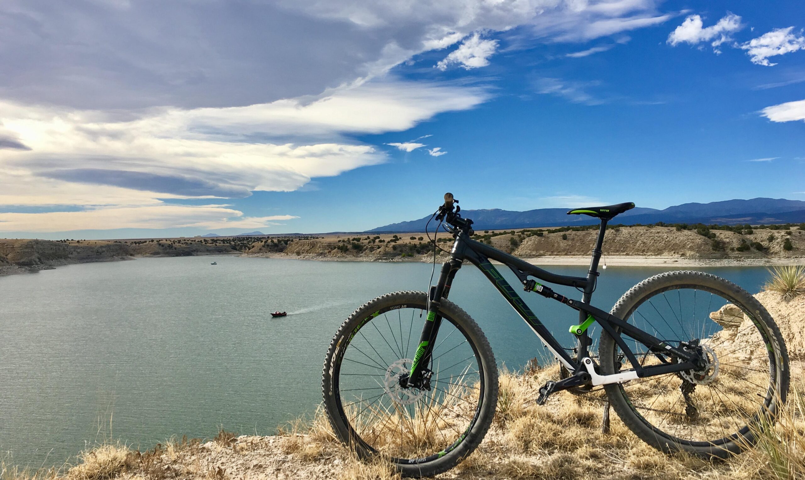 Fuji Rakan: A mountain bike rests on a grassy cliff overlooking a serene lake surrounded by hills and mountains under a bright blue sky with scattered clouds. In the distance, a small boat is visible on the water.