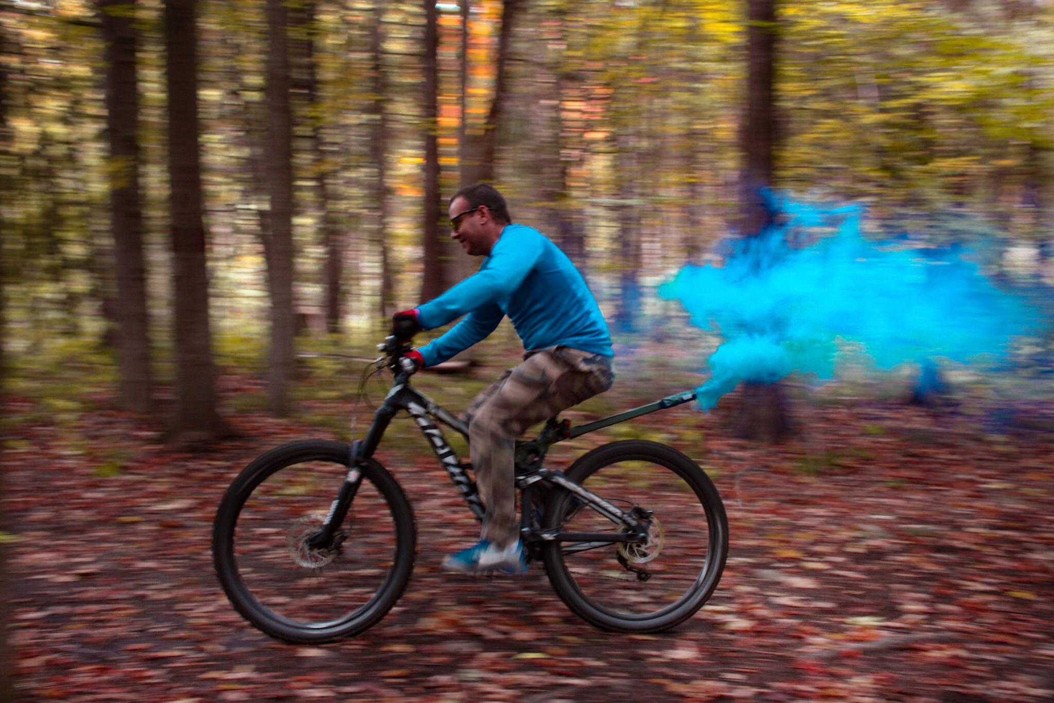 Devinci Troy: A cyclist riding a mountain bike through a colorful autumn forest, with blue smoke trailing behind as he speeds along a path covered in fallen leaves. The scene captures a sense of motion and adventure in a vibrant natural setting.