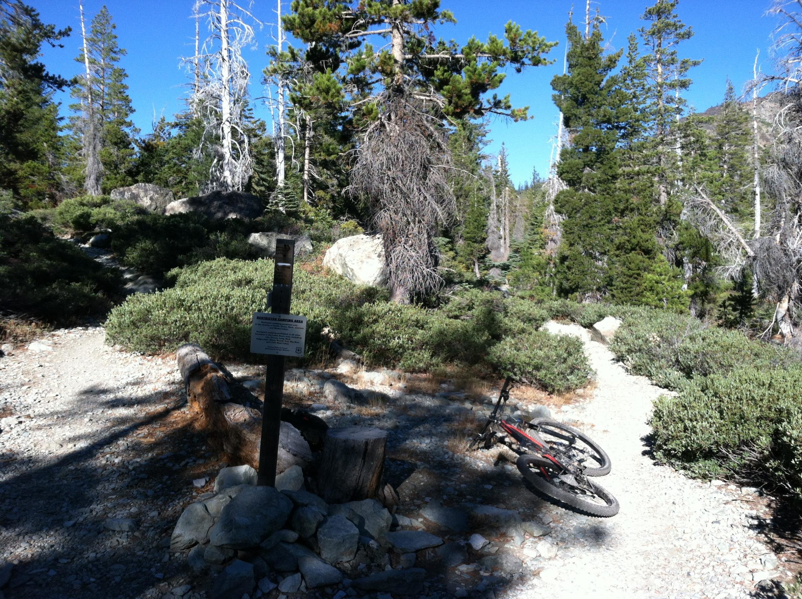 A mountain bike rests on the ground near a trail junction surrounded by lush greenery and rocky terrain in a forested area. A signpost nearby indicates the location of a designated camping area. The sky is clear and blue, showcasing a bright, sunny day. Long Lake mountain bike trail.