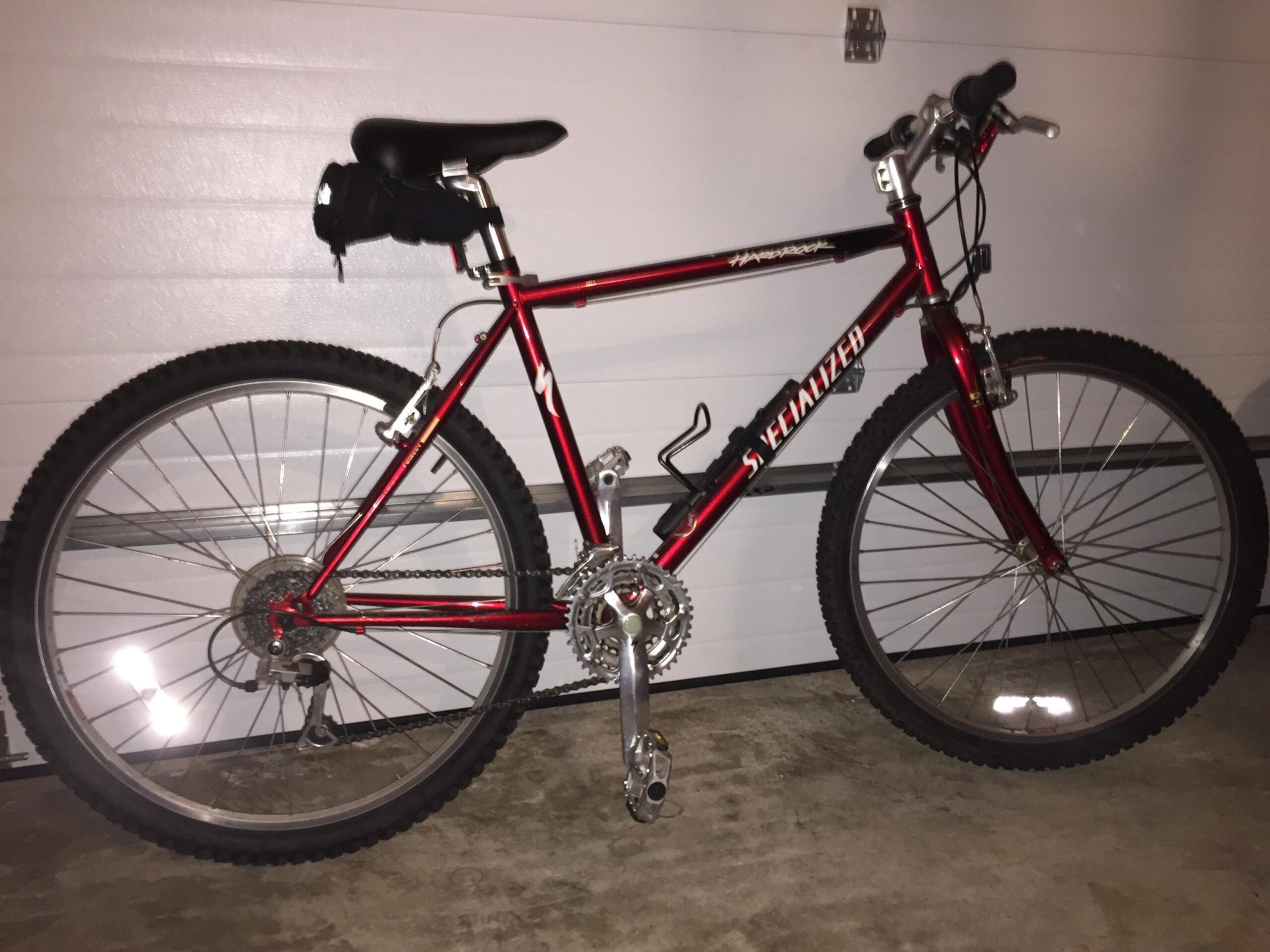 Specialized hardrock: A red mountain bike with thick tires and a black seat, parked against a garage wall. The frame is sleek with visible branding, and it features a gear system and pedals. The garage floor is concrete, adding to the indoor setting.